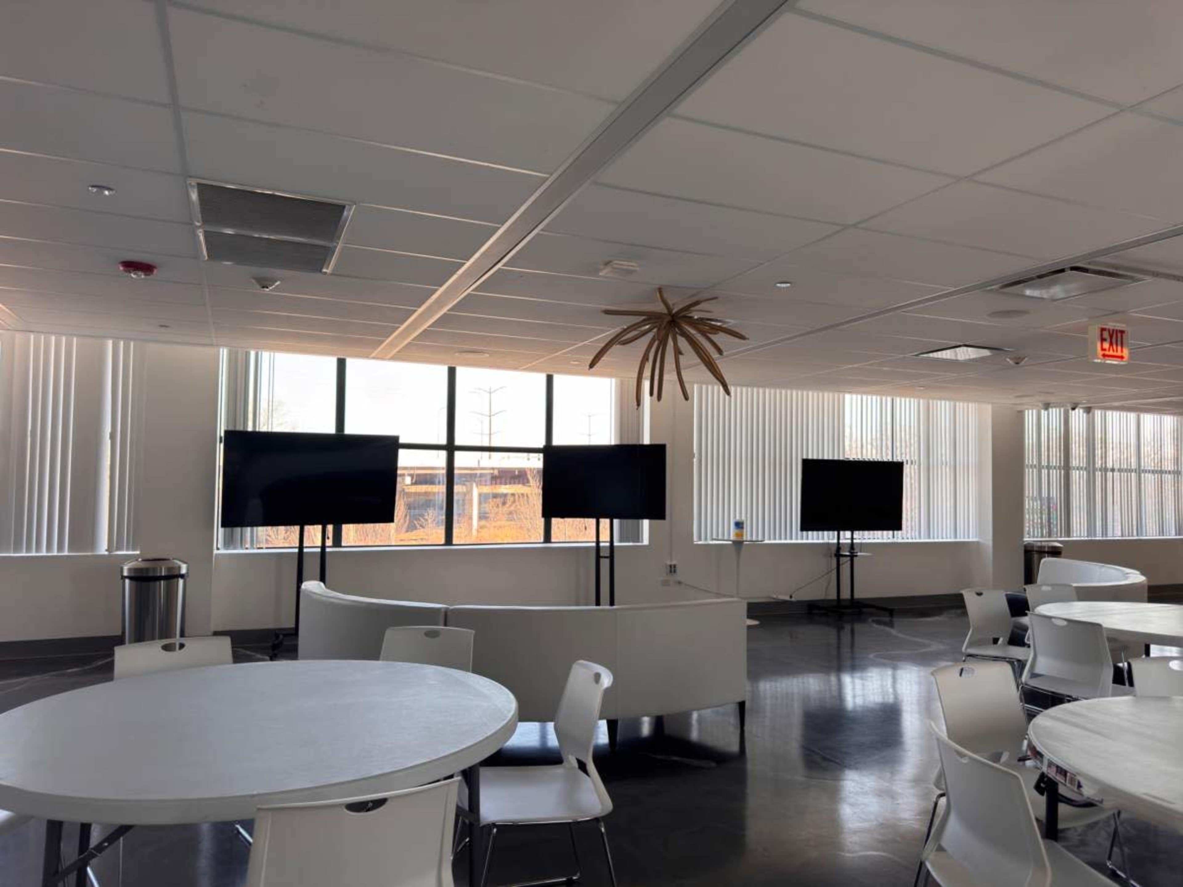 The image shows a modern room with two large screens on stands, circular tables with chairs, and a decorative element hanging from the ceiling.