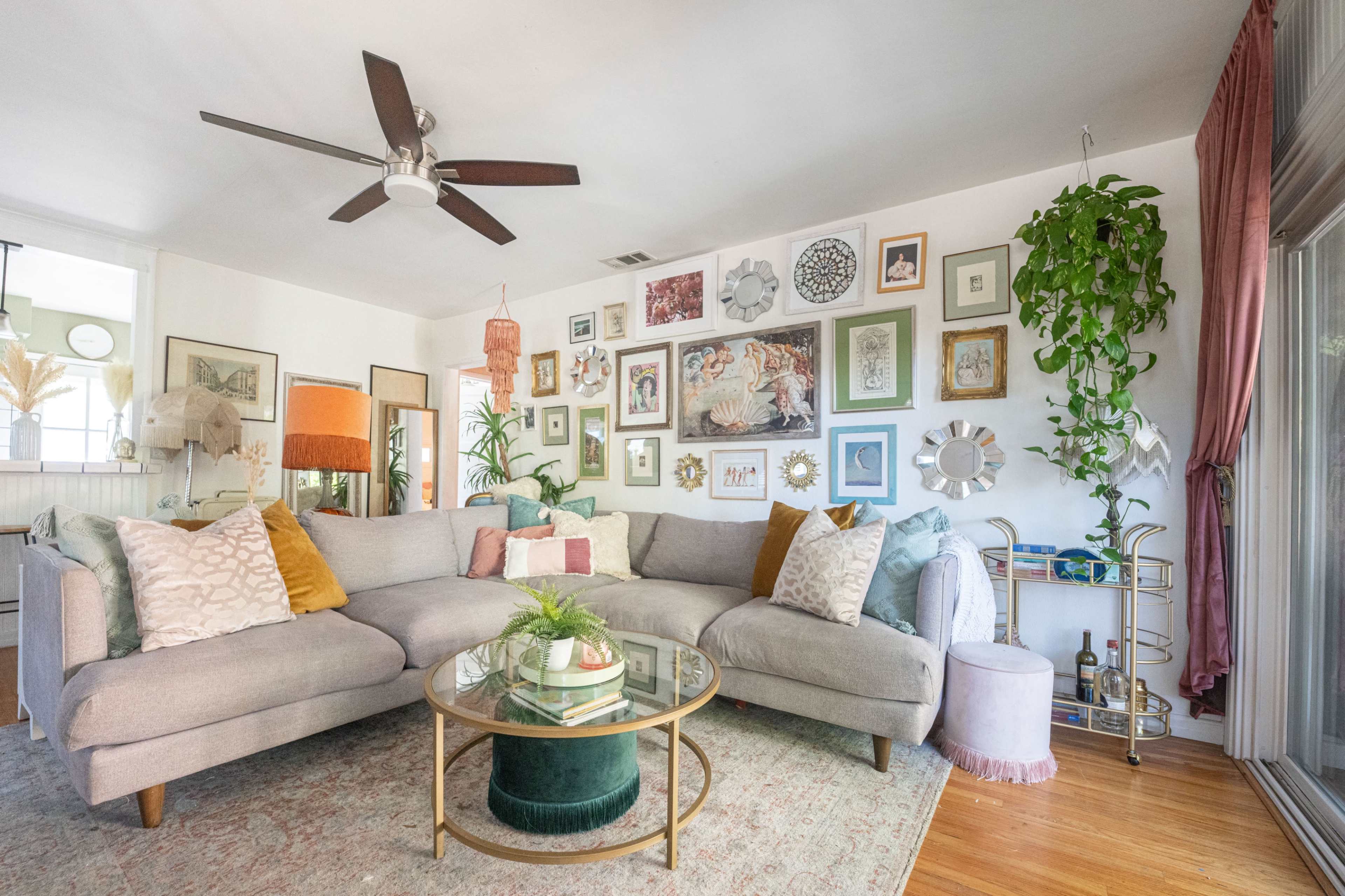 A light-filled living room features a large gray sectional sofa with decorative pillows, a round glass coffee table, and an art-covered wall adorned with various framed pictures and plants.