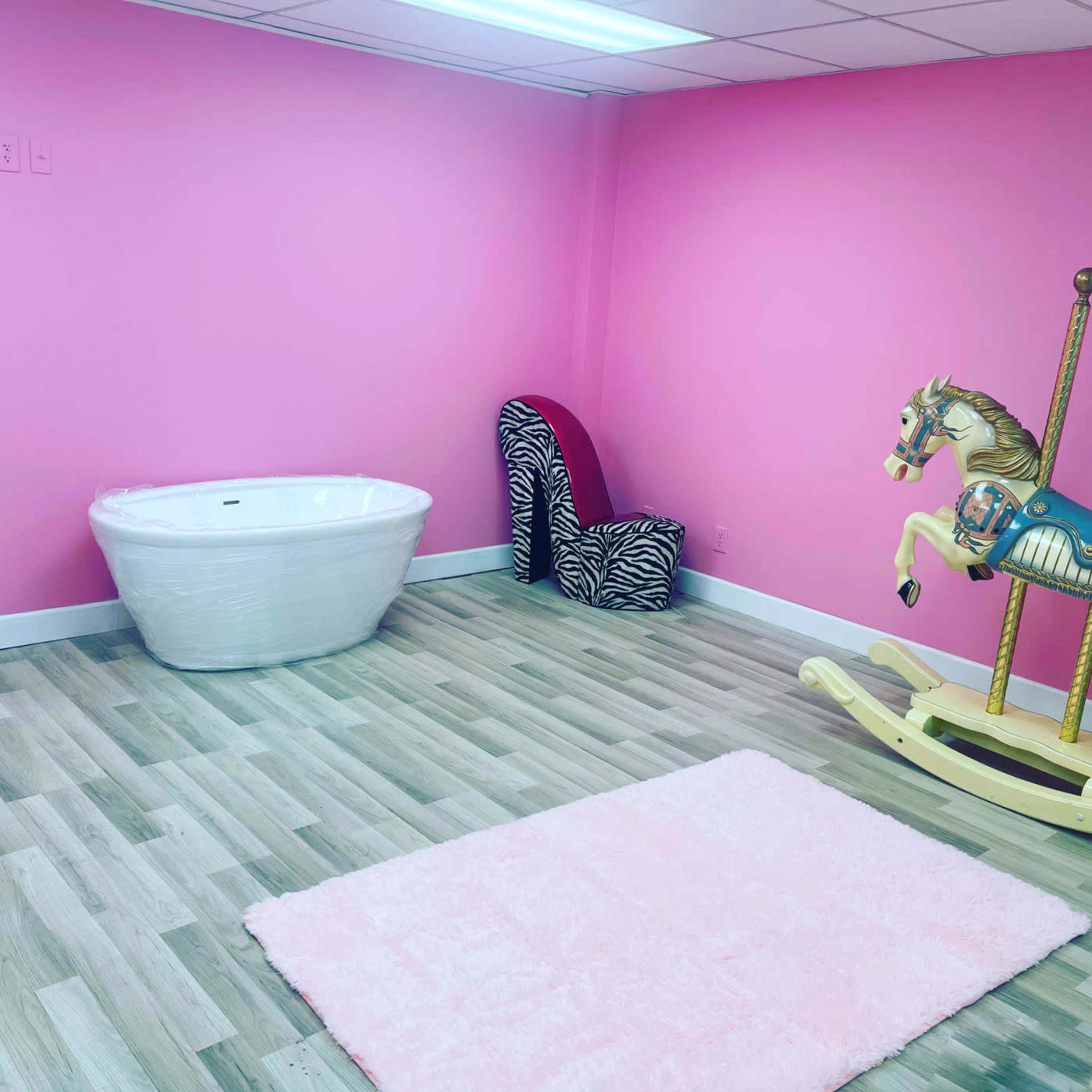 The room features pink walls, a white freestanding bathtub, a zebra-patterned chair, a wooden rocking horse, and a light pink rug on a wooden floor.