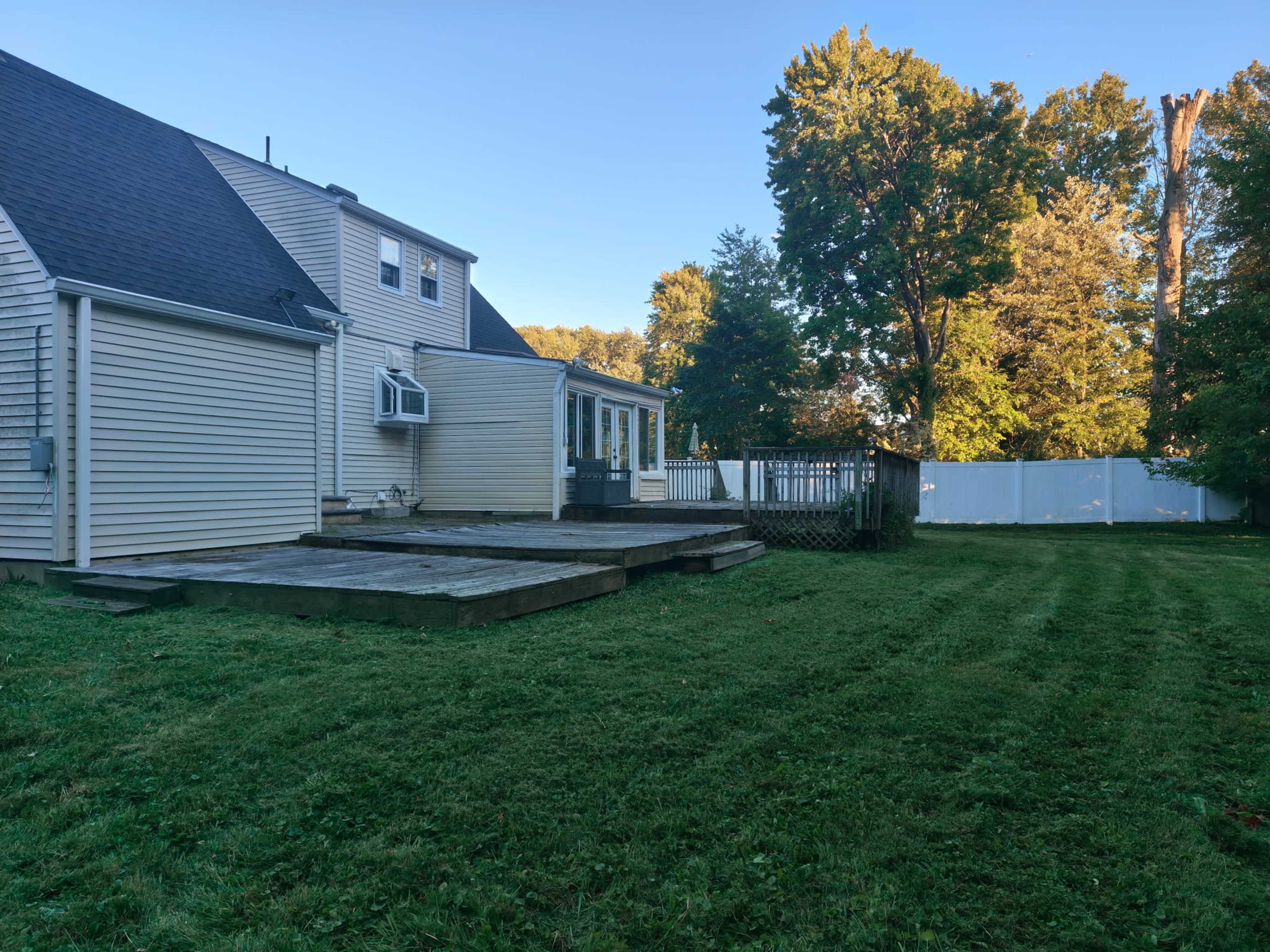 The image shows a backyard with a wooden deck adjacent to a house, surrounded by grass and a white fence in the background.