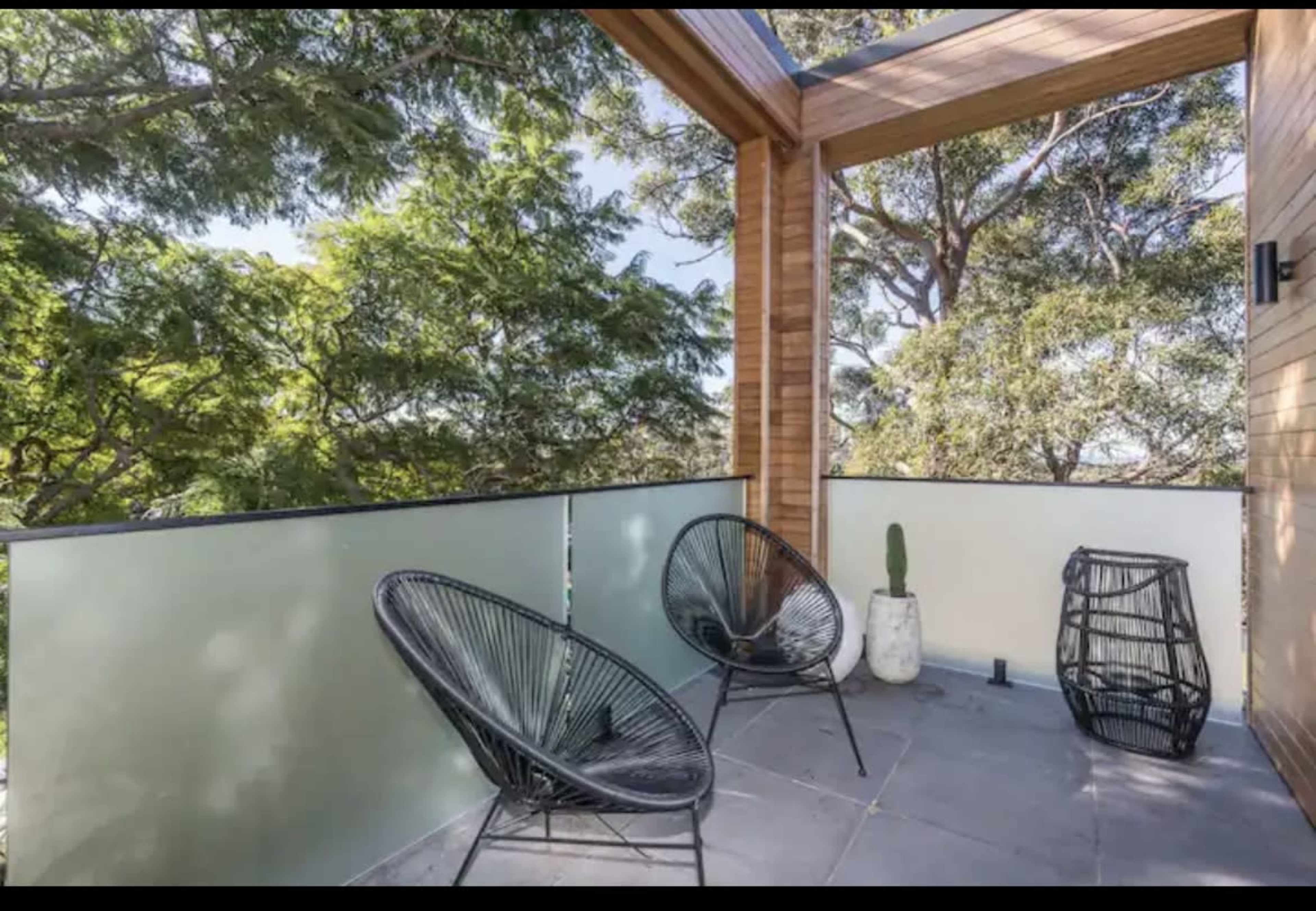 The image shows a modern balcony with two black chairs, a potted cactus, and a decorative basket, surrounded by greenery.