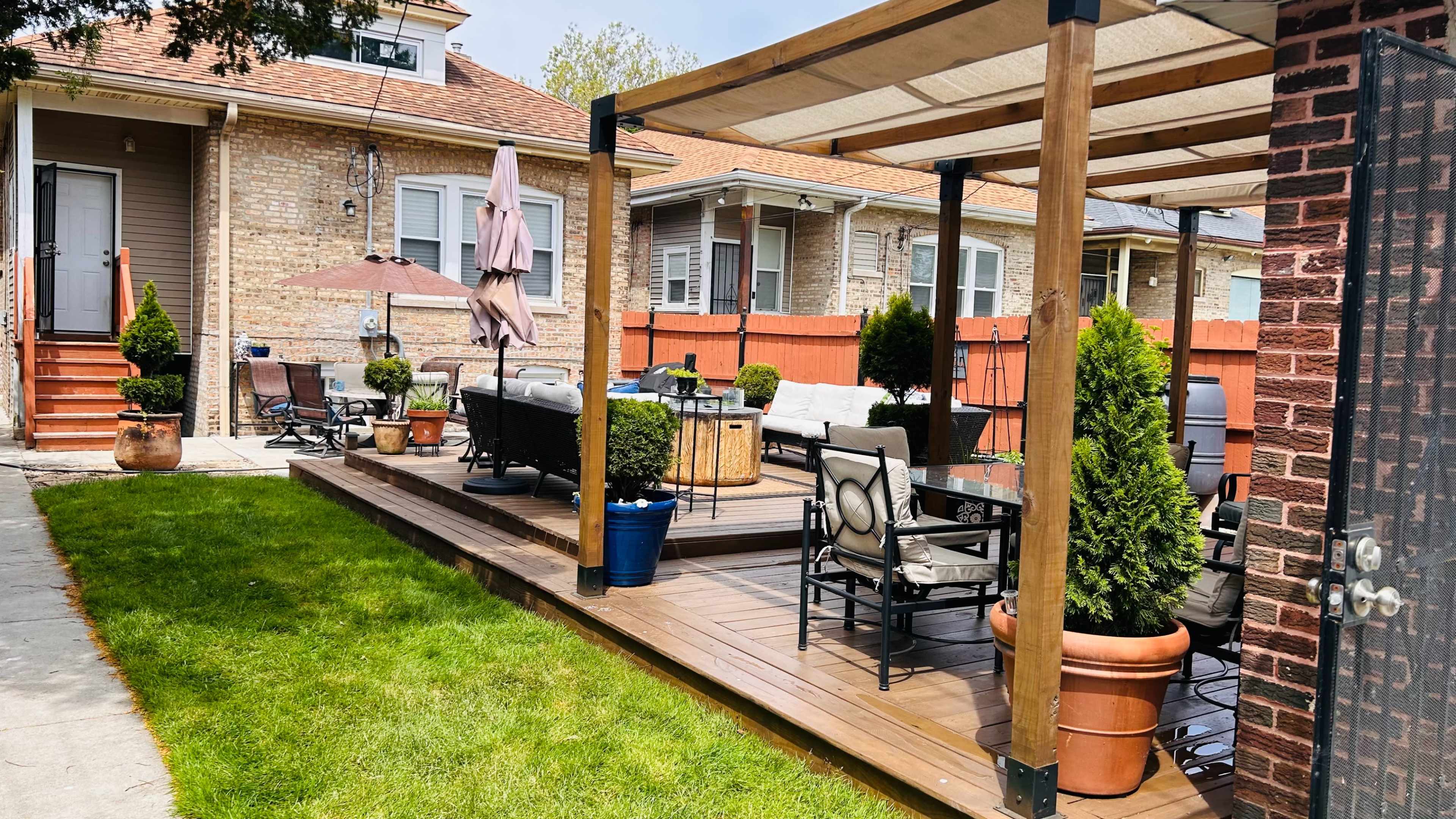 The image shows a wooden patio area with seating arrangements, potted plants, and a pergola, adjacent to a brick building and a grassy lawn.