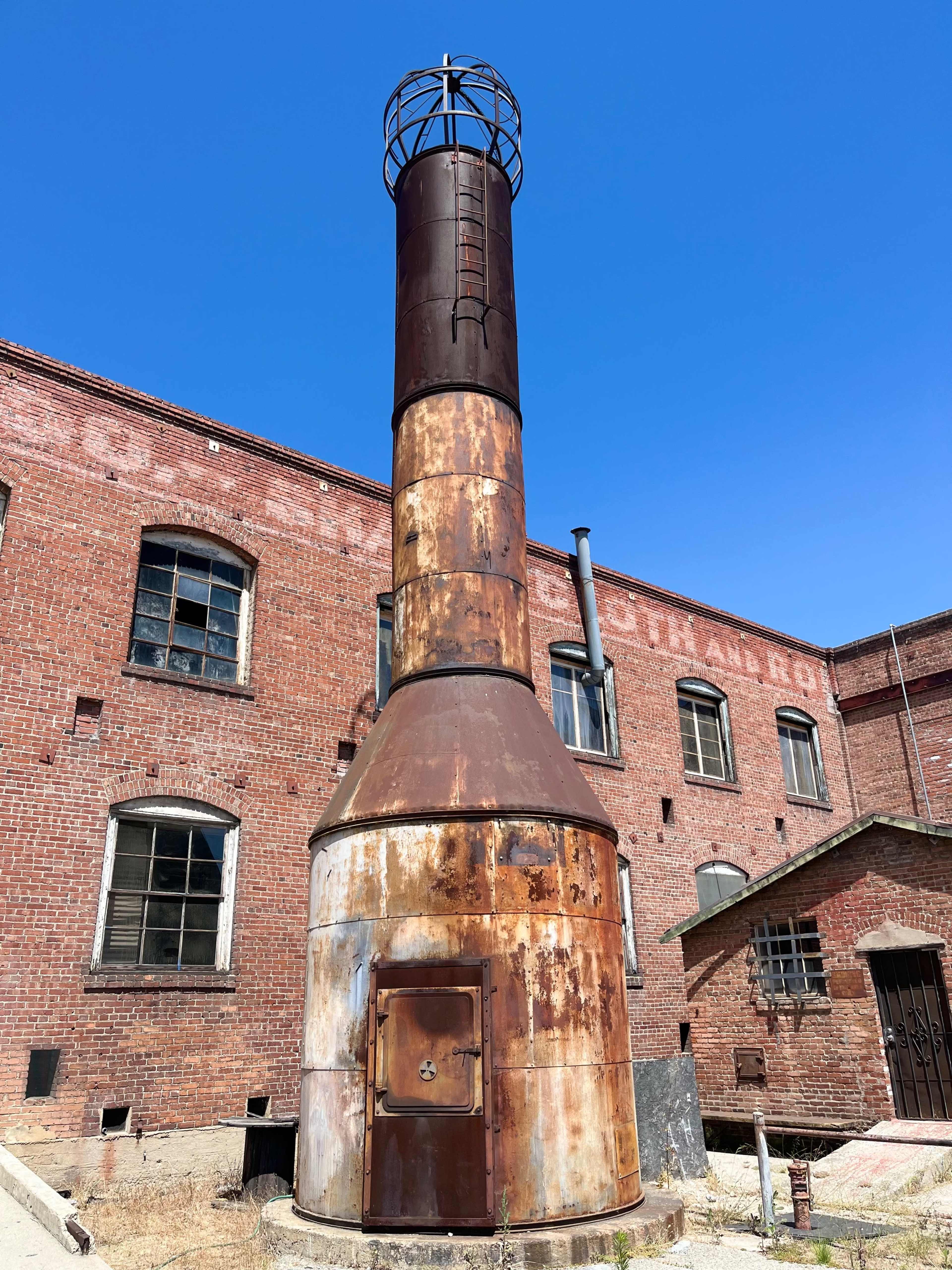 A tall, rusted industrial chimney stands next to a brick building under a clear blue sky.