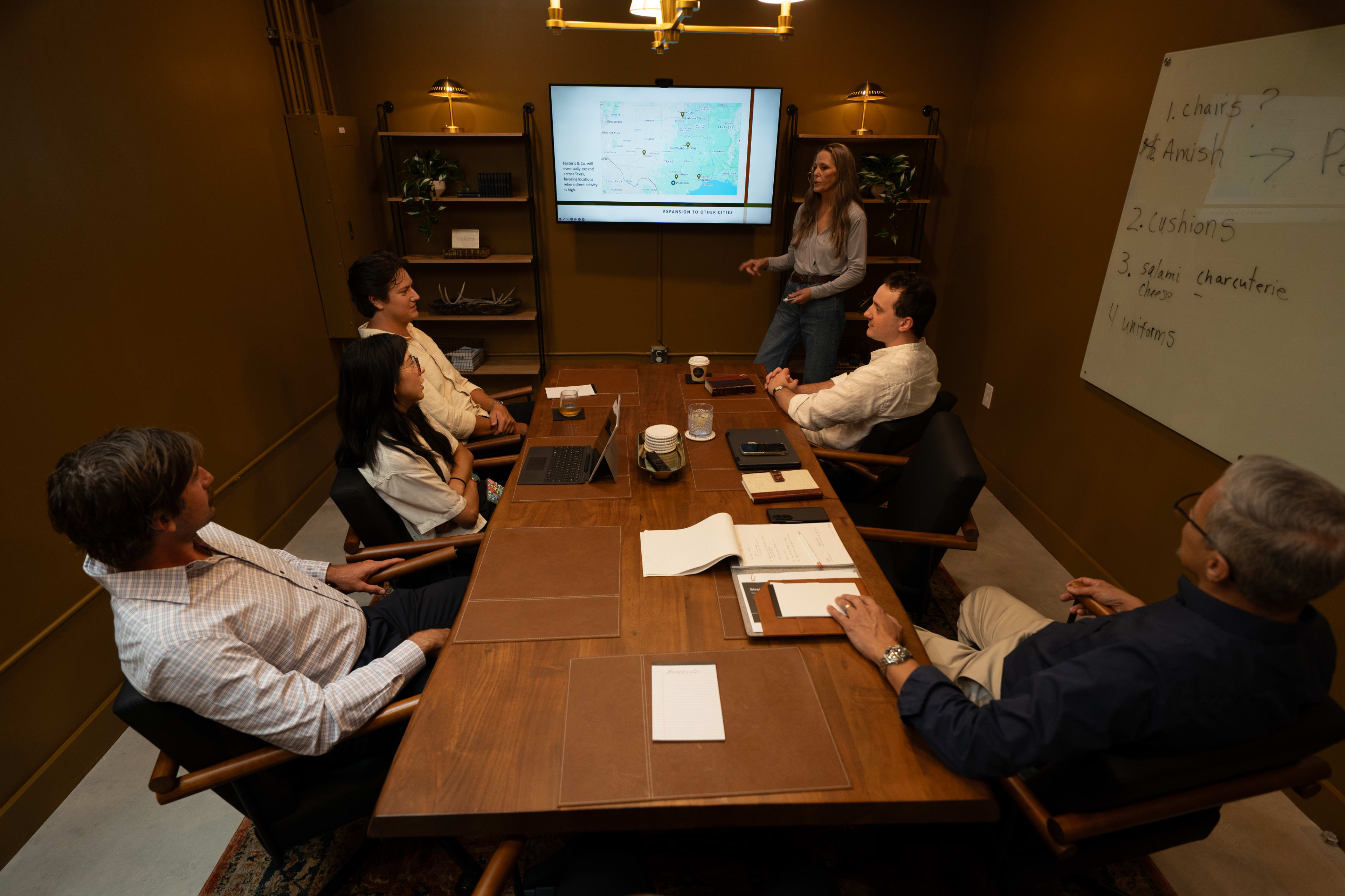 A group of six people is seated around a conference table, with one person presenting a map on a screen in a well-lit meeting room.