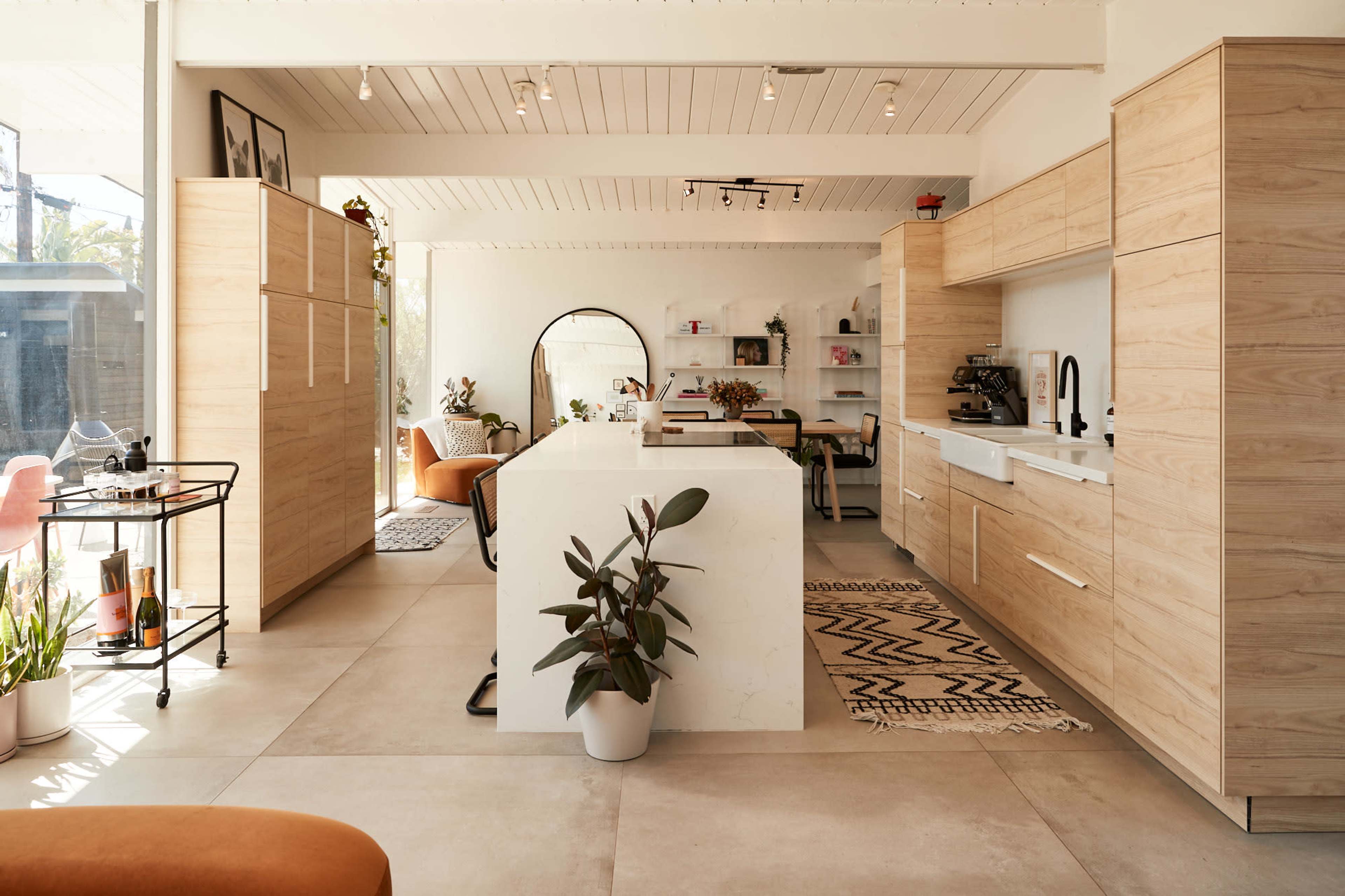 A modern kitchen featuring light wooden cabinetry, a central island, and a variety of indoor plants in a spacious, well-lit room.