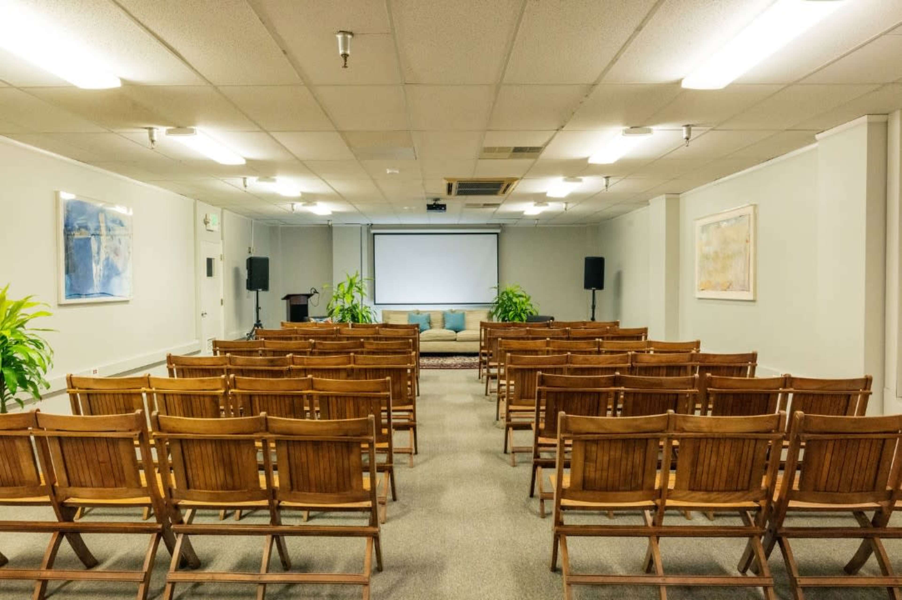 A semi-filled conference room with wooden chairs arranged in rows facing a large screen and a seating area at the front.