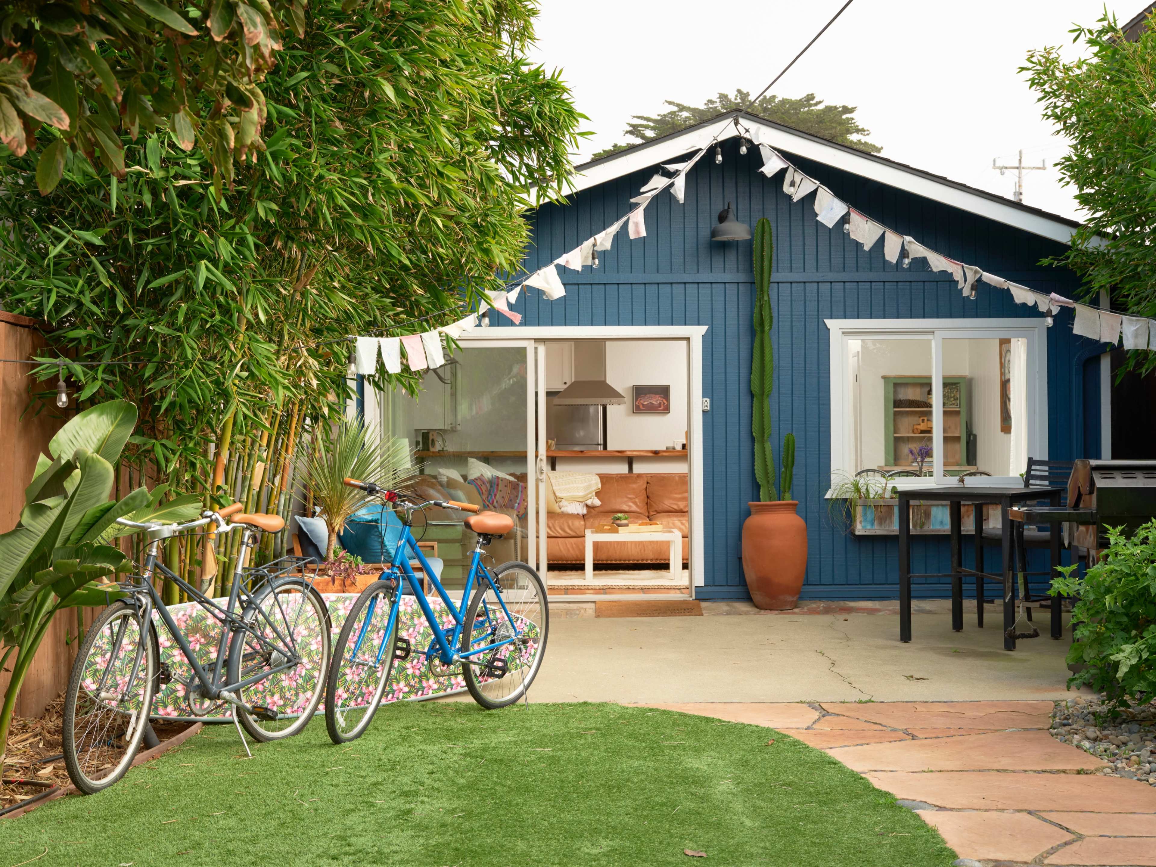 A blue cottage with an open front and a patio is surrounded by greenery and two bicycles are parked nearby.