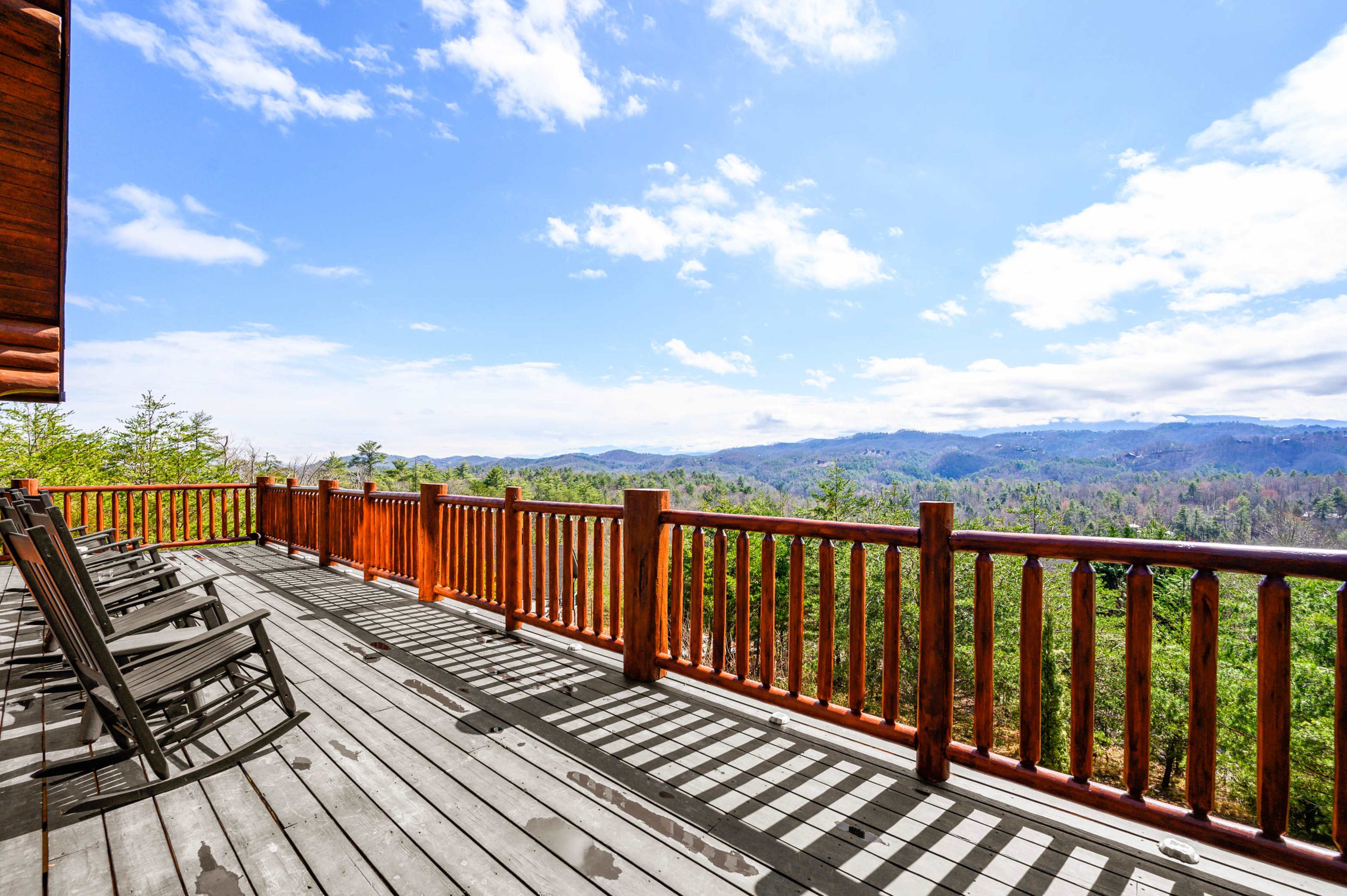 A wooden deck features rocking chairs and overlooks a mountainous landscape under a partly cloudy sky.