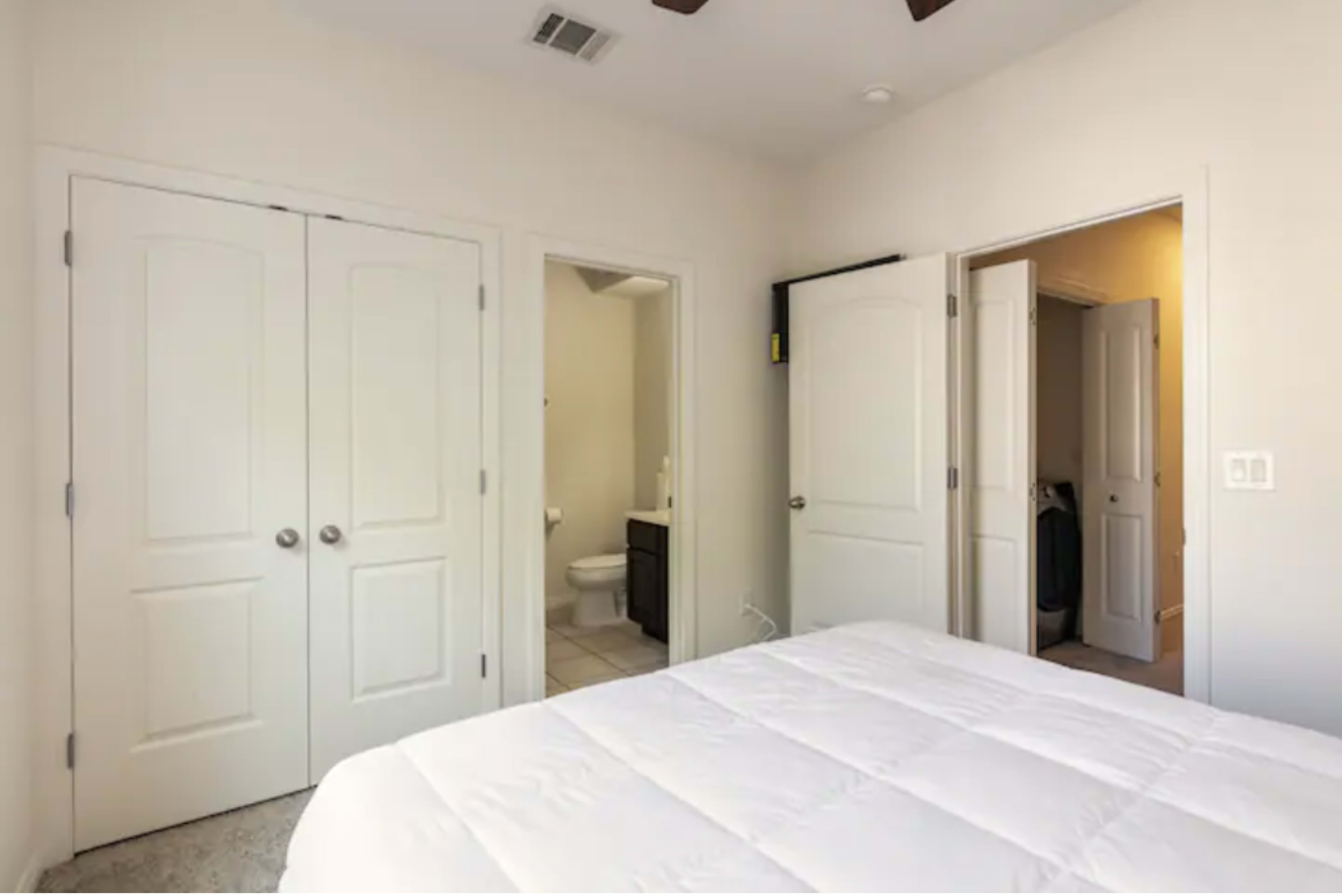 The image shows a bedroom with a bed in the foreground and two open doorways leading to other rooms, one of which is a bathroom.