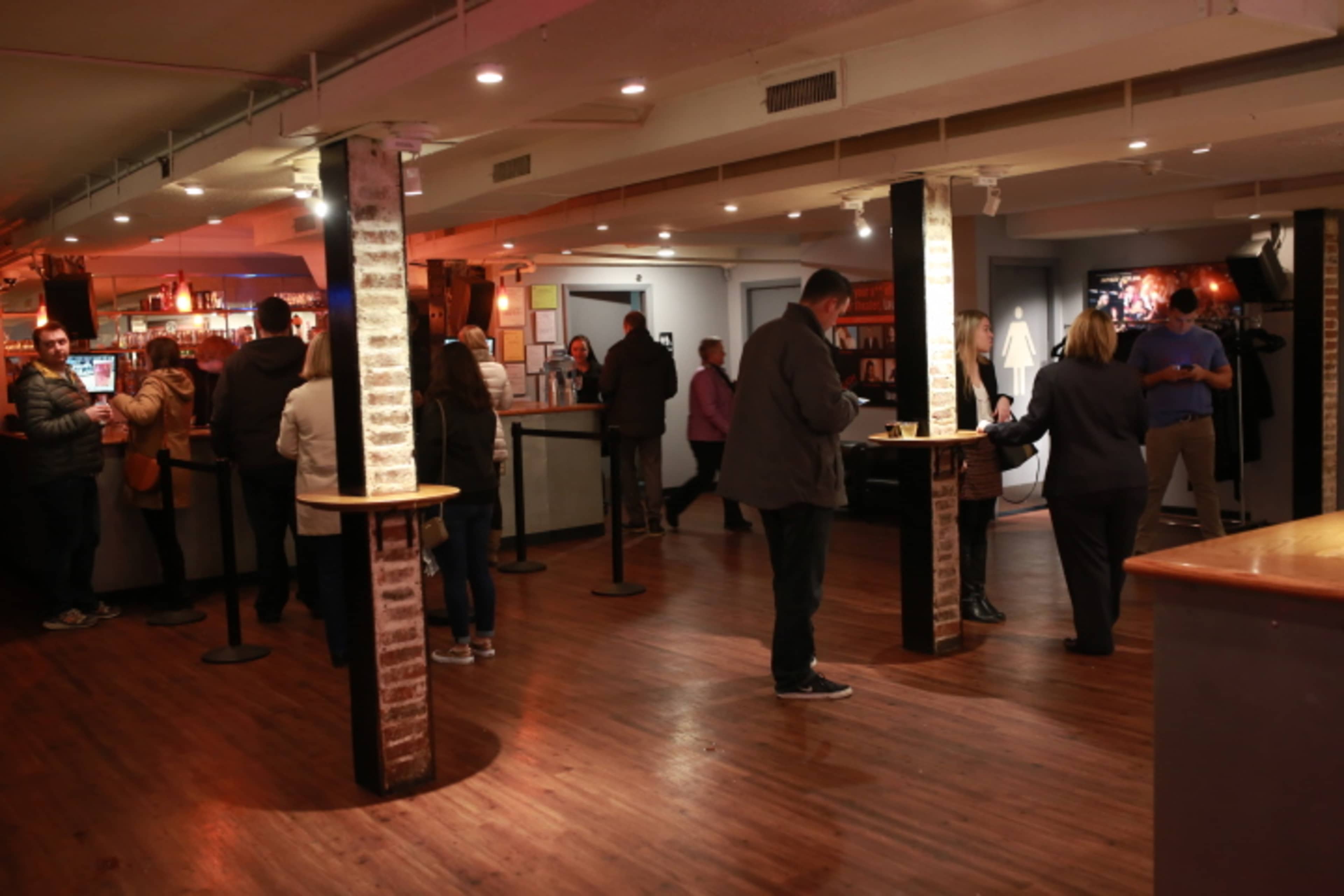A group of people socialize and order drinks in a dimly lit bar with a wooden floor and exposed brick pillars.