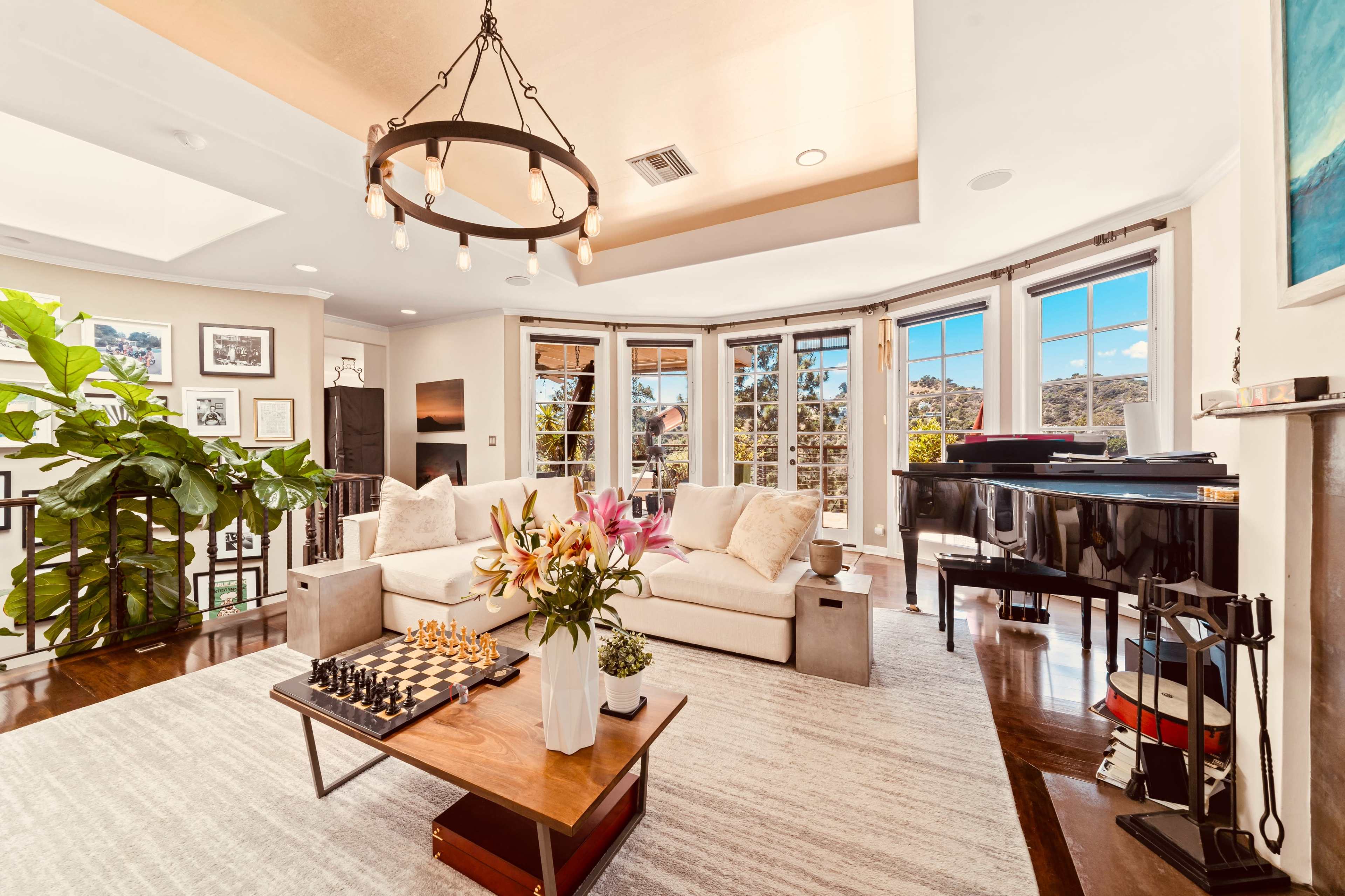 The image shows a living room with large windows, a beige couch, a grand piano, and a coffee table with flowers and a chess set.