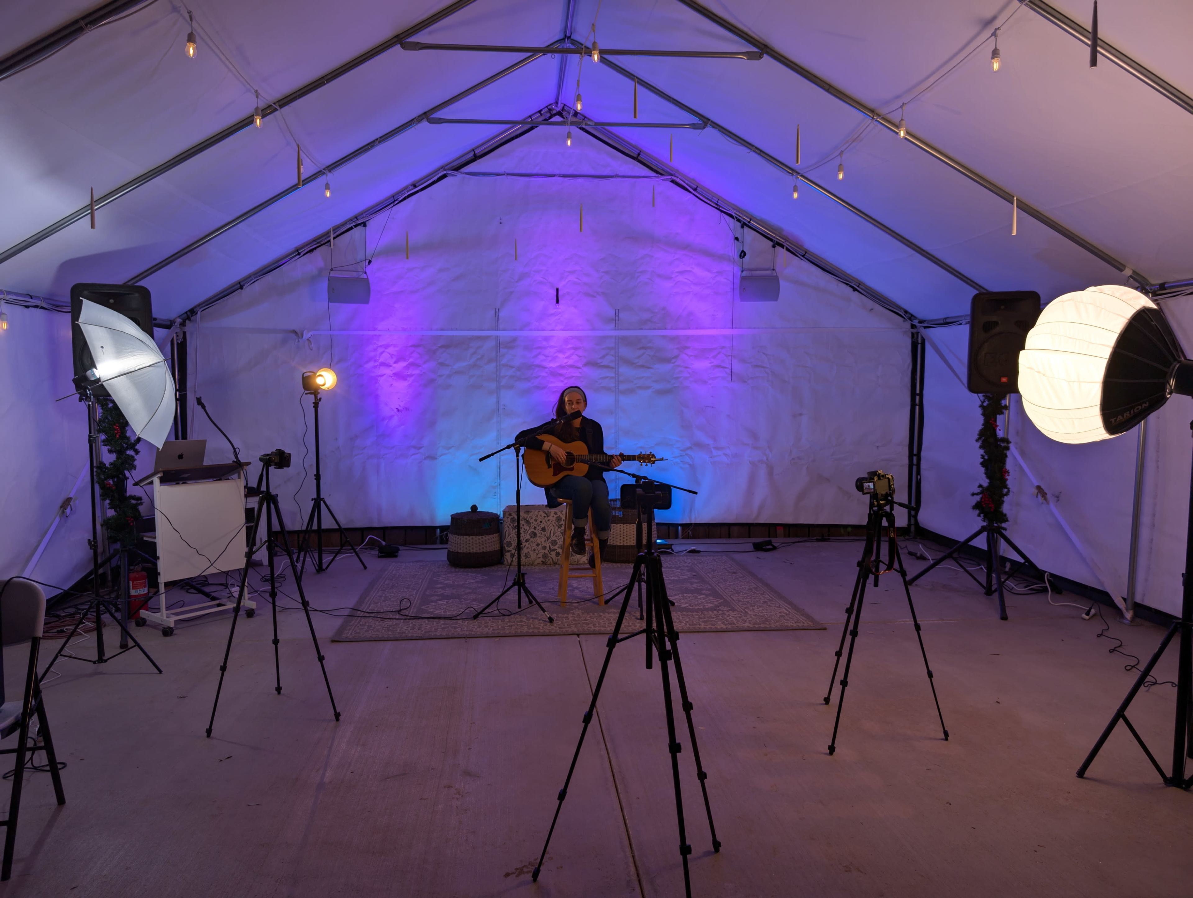 A musician plays guitar in a dimly lit tent surrounded by cameras and studio lighting.