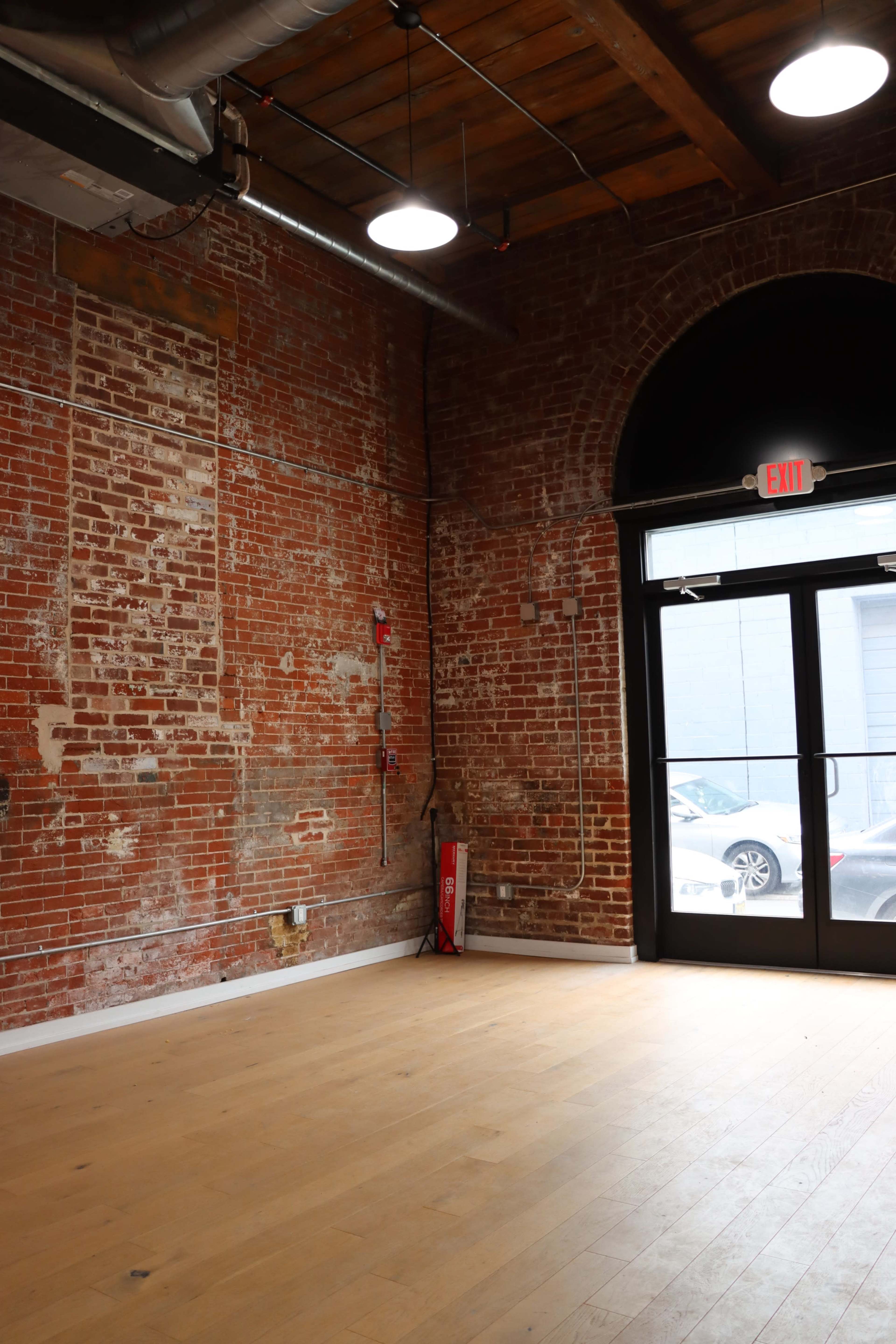 The image shows an empty room with exposed brick walls, wooden flooring, and large windows with a door leading outside.