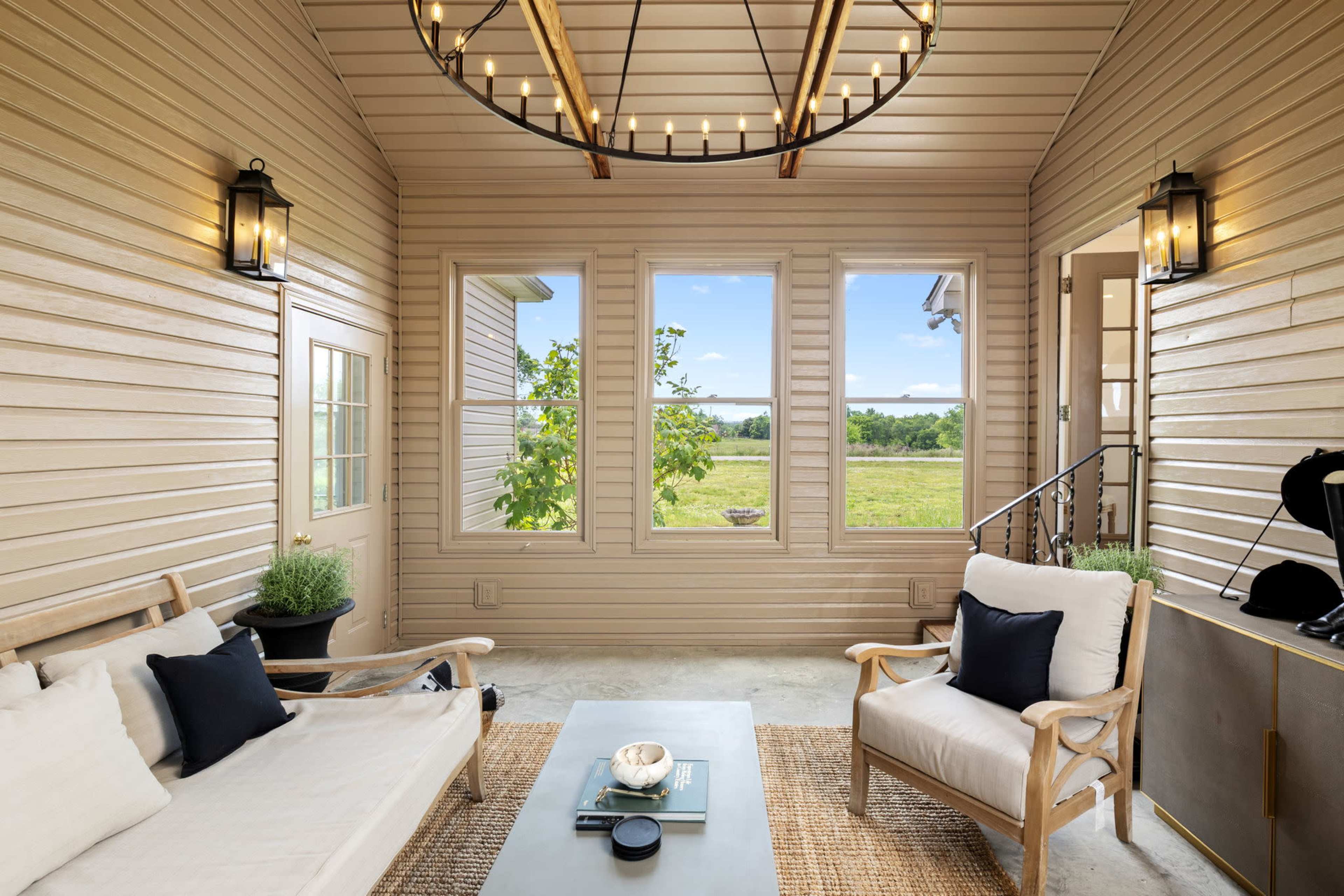 The image shows a cozy porch with a wooden sofa and armchair, a coffee table, and large windows overlooking a green landscape.
