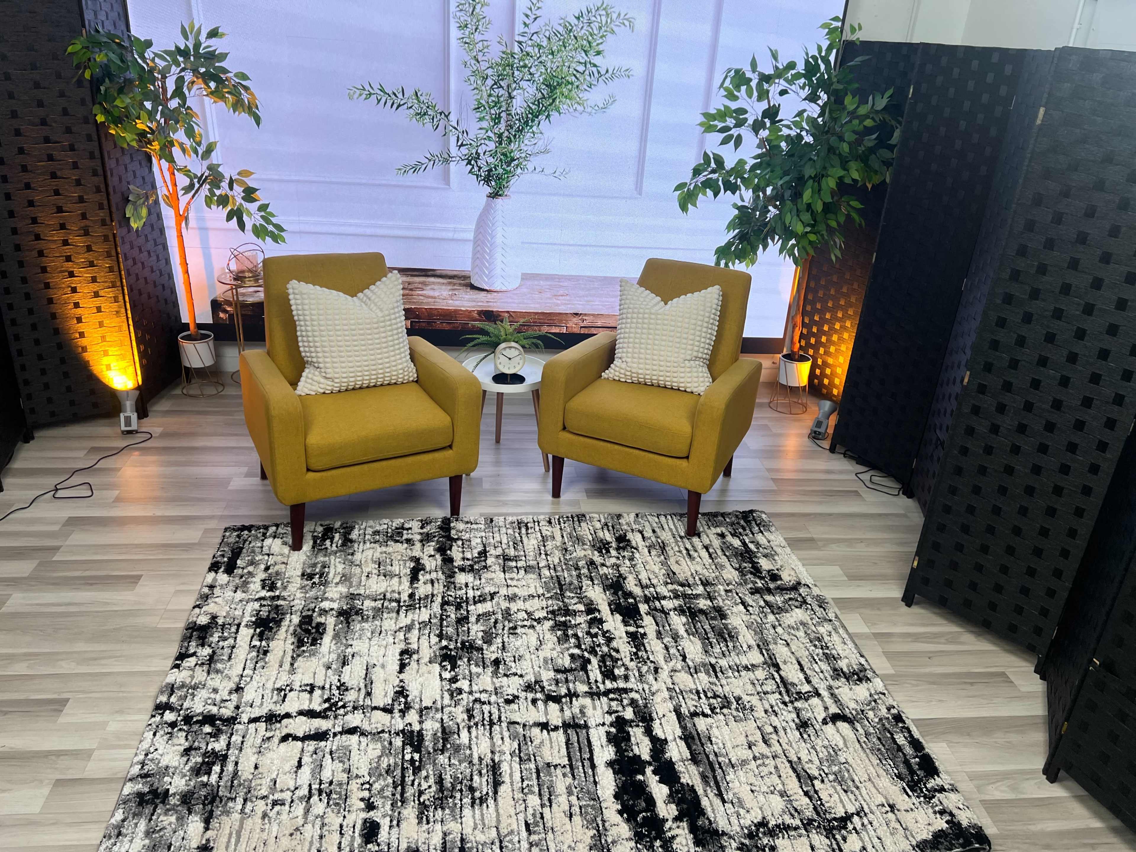 The image shows two yellow armchairs with decorative pillows facing each other on a patterned rug, surrounded by plants and a backdrop featuring a large screen with a decorative arrangement.