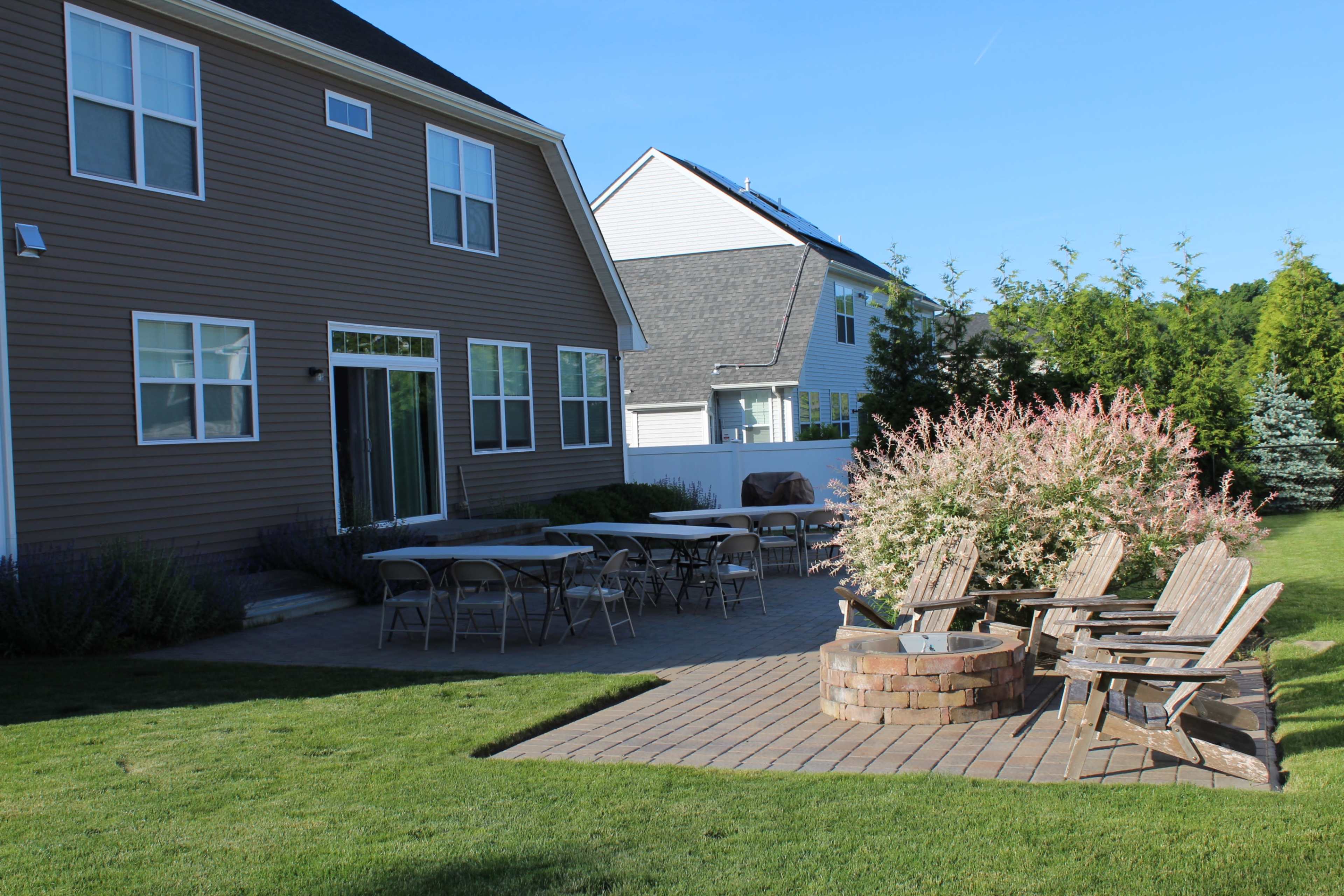 A backyard patio area with several tables and wooden chairs arranged around a circular stone fire pit, framed by green grass and shrubs.