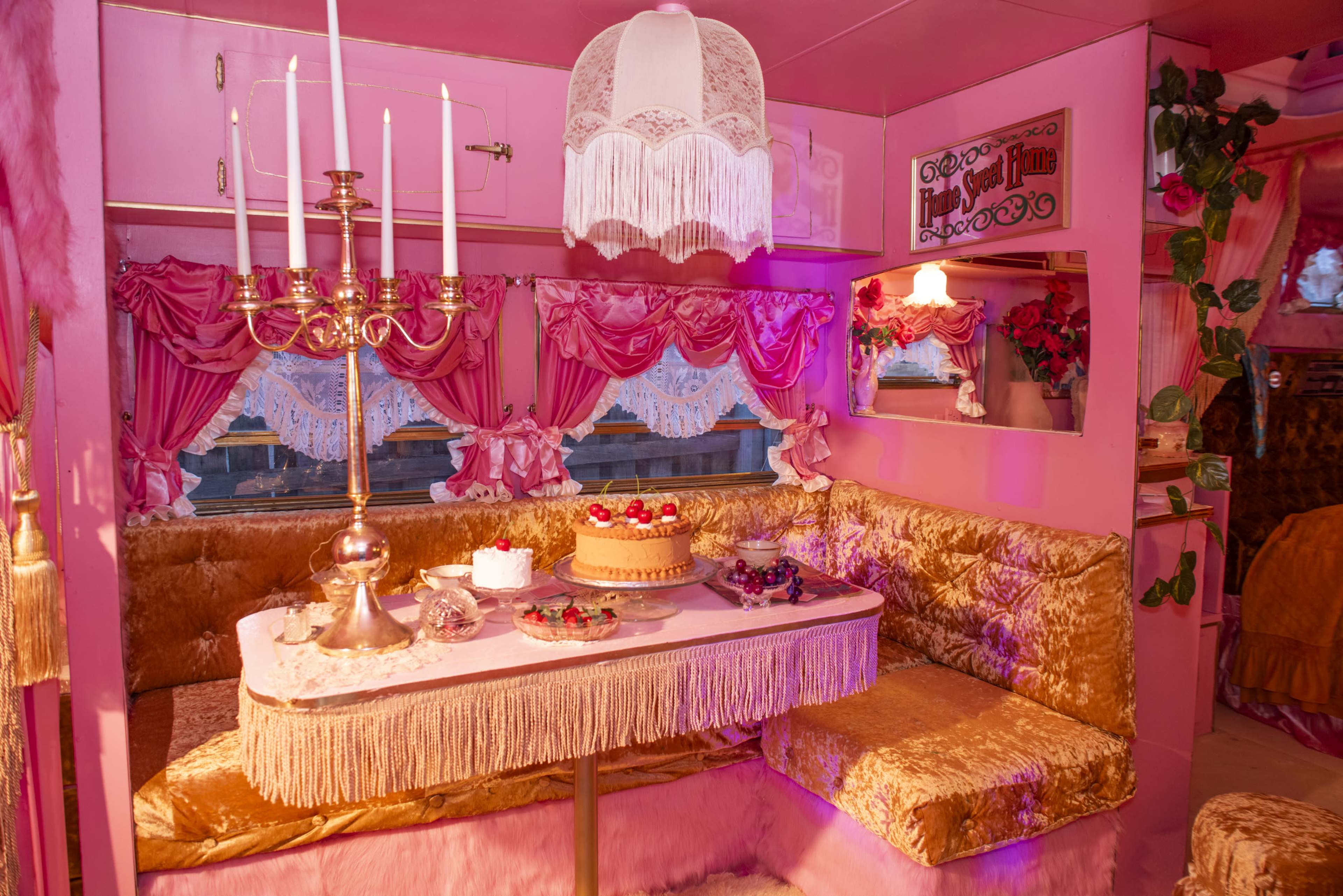 The interior of a pink-themed vintage trailer features a dining area with a lace chandelier, plush seating, and a table set with a cake and decorations.