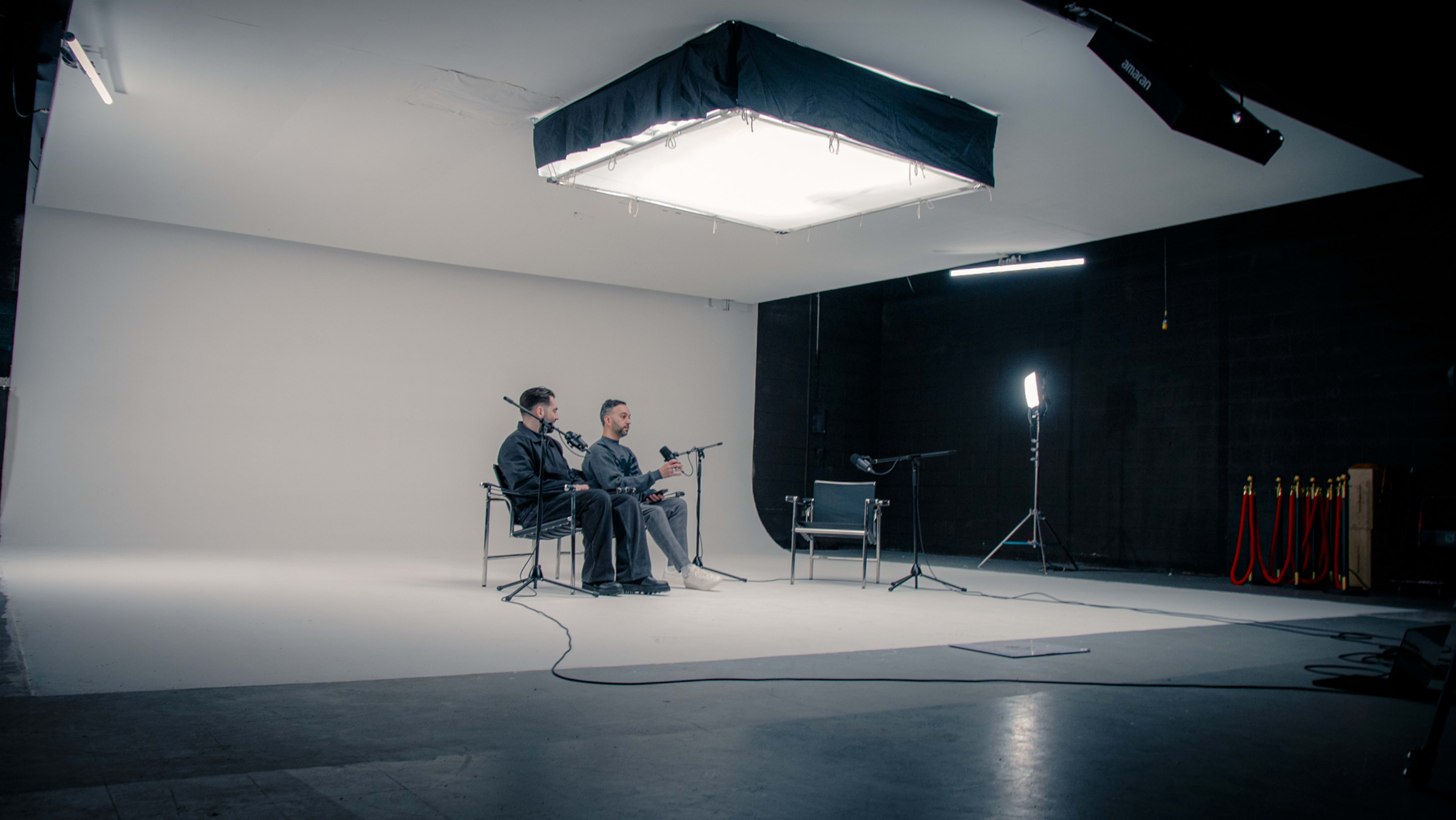 Two men sit on chairs in a minimalist studio setting, with a large softbox light overhead and a microphone setup nearby.