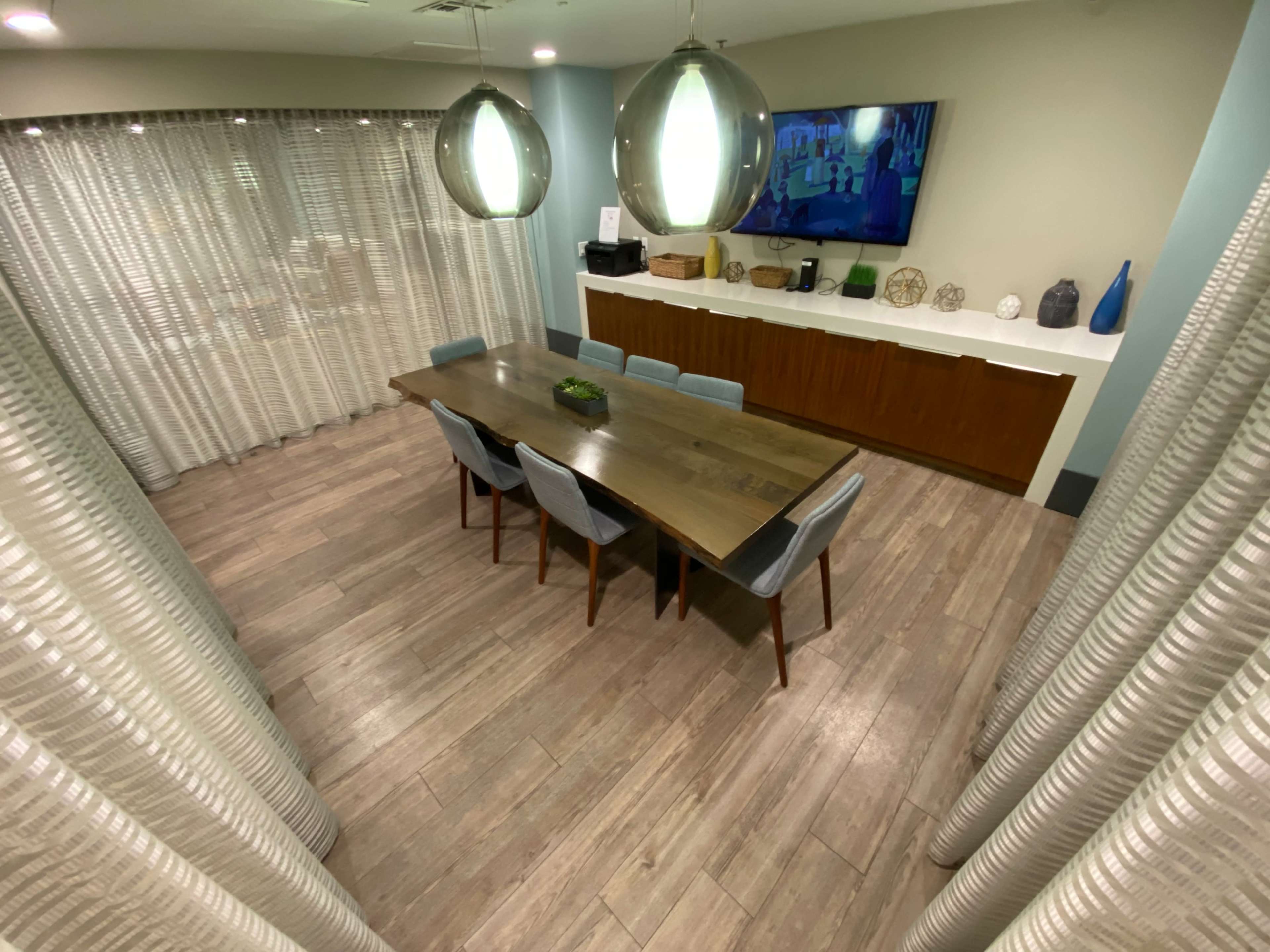 A spacious conference room features a long wooden table surrounded by six blue chairs, with a television mounted on the wall and sheer curtains on the windows.