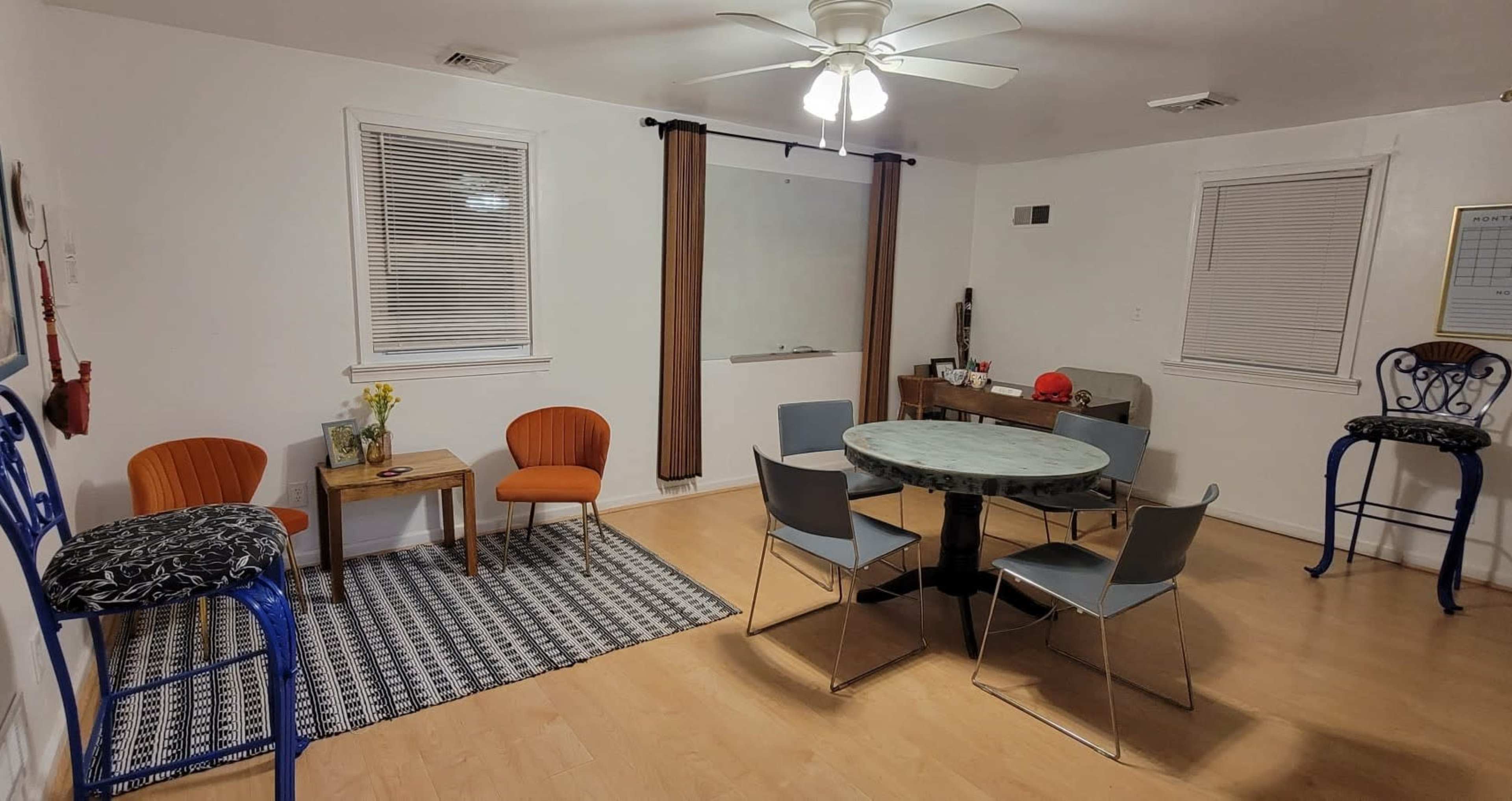 A sparsely furnished room featuring a round table with chairs, two orange chairs, and a patterned rug on a wooden floor.