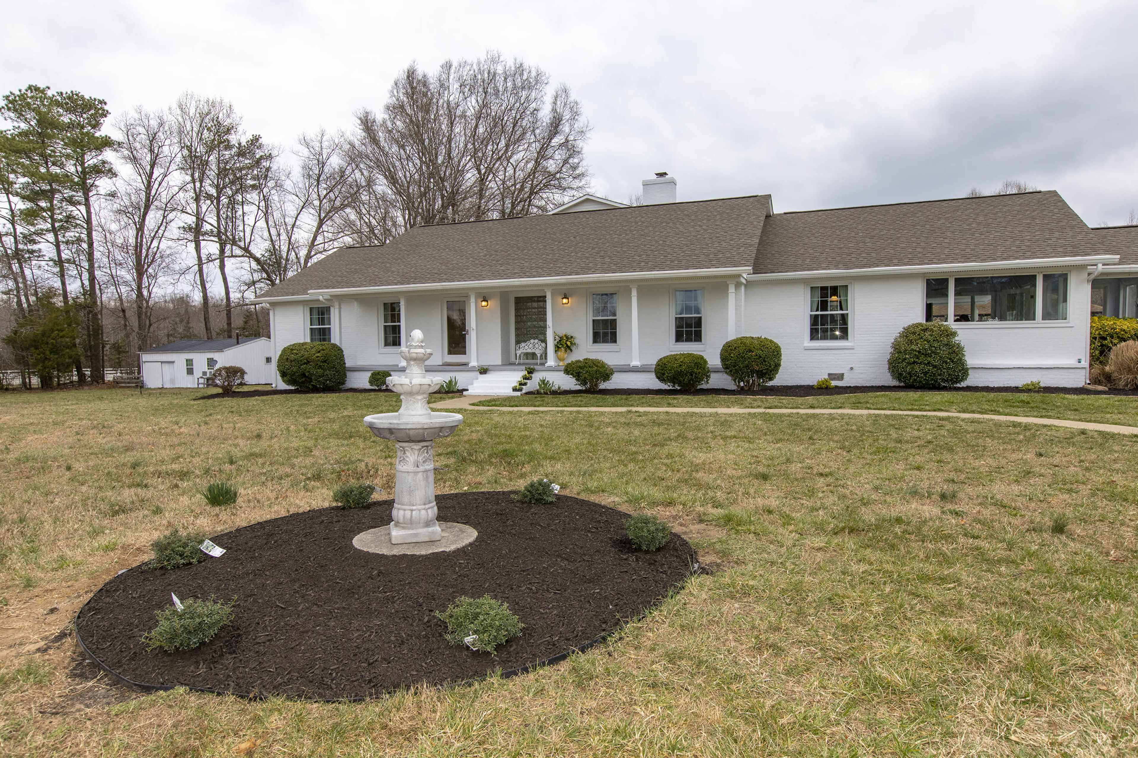 A white house with a gray roof features a central fountain surrounded by a circular flower bed and well-manicured bushes in the front yard.
