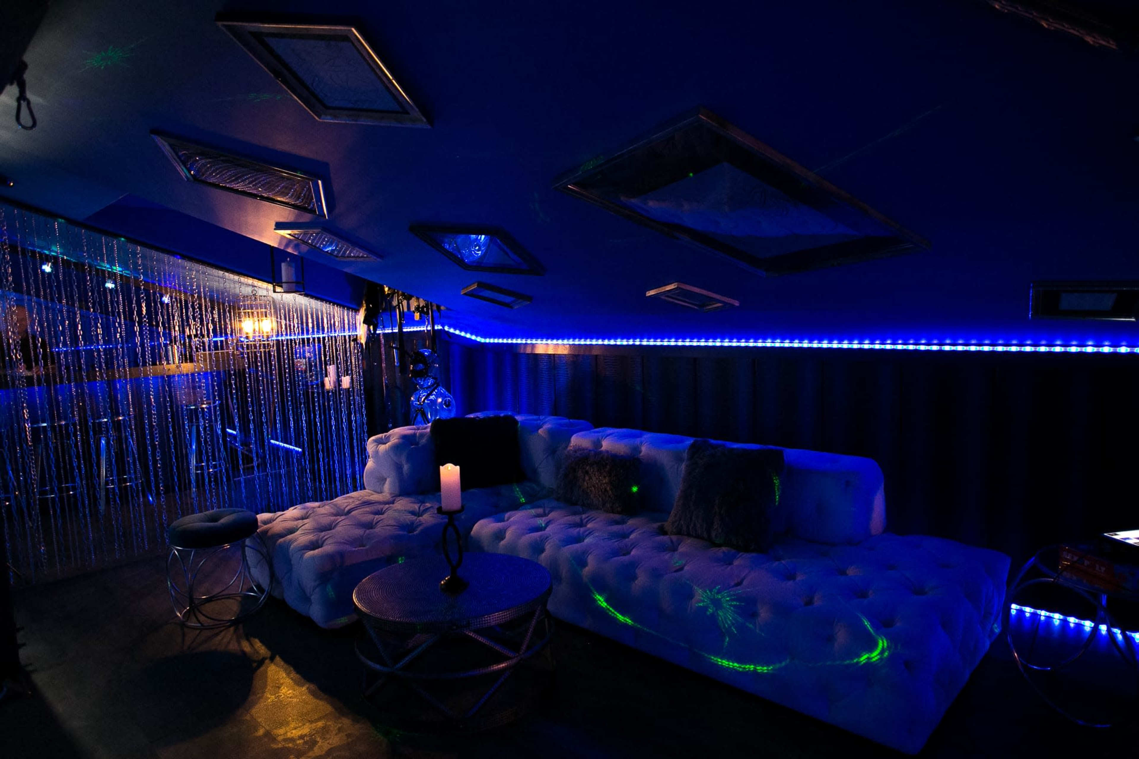 A dimly lit lounge area featuring a white tufted sectional sofa, a circular coffee table, and blue LED lighting along the walls.