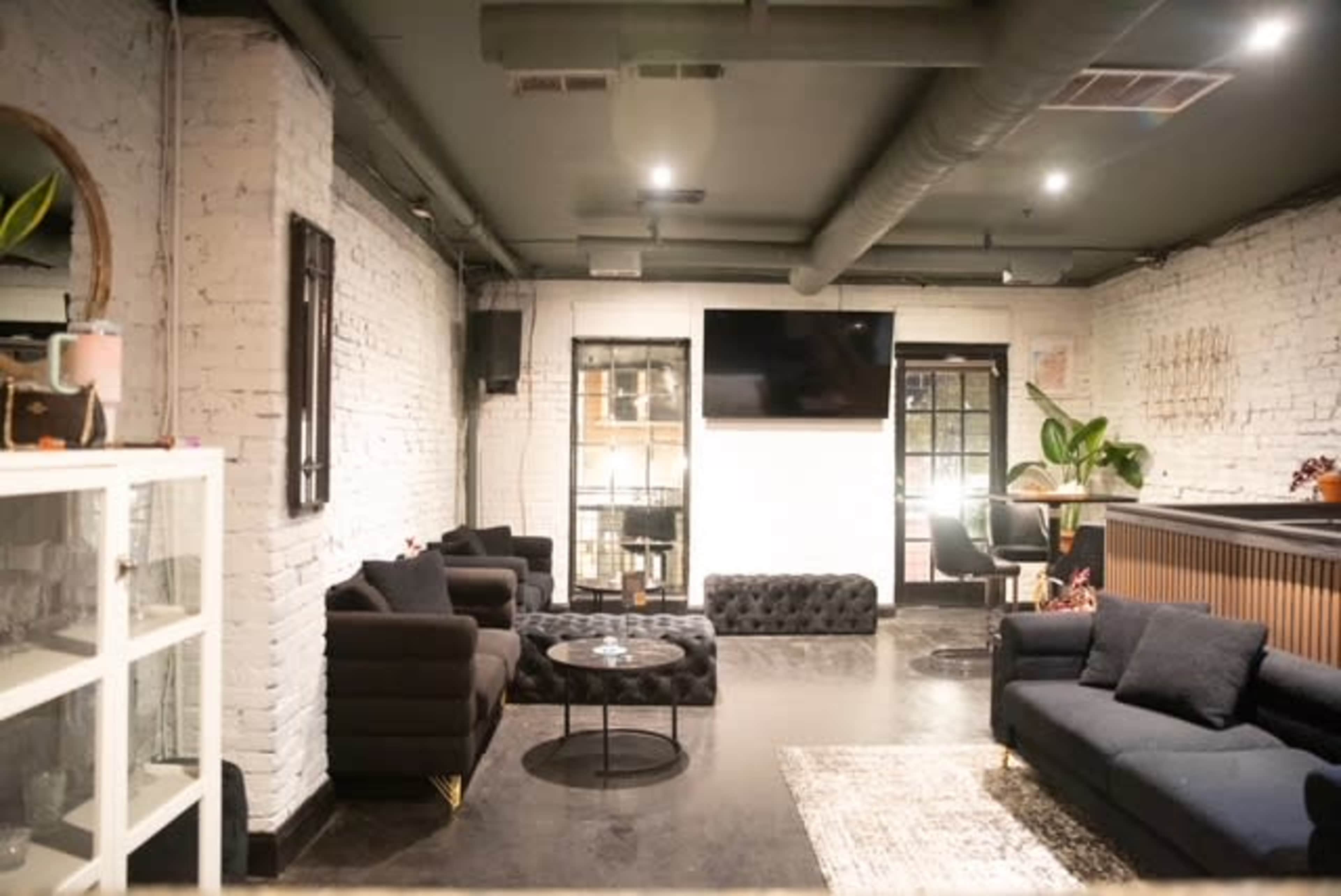 The image shows a modern lounge area with dark furniture, a flat-screen TV mounted on a white wall, and large windows.