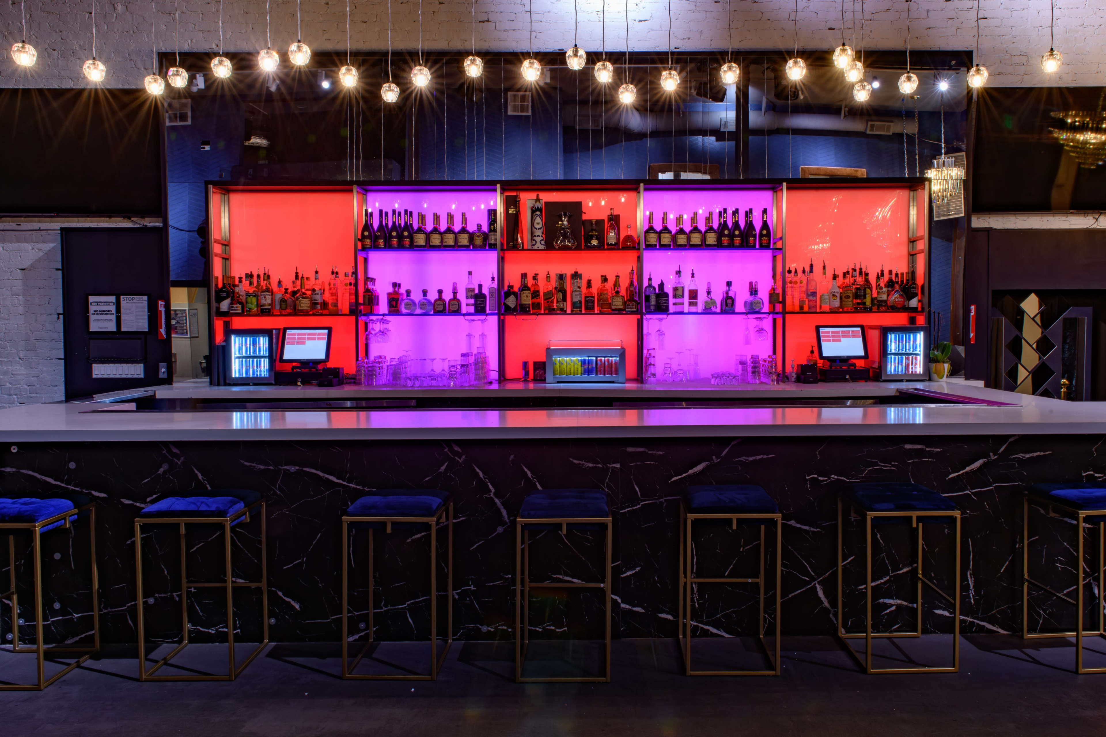 The image shows a modern bar with illuminated shelves showcasing various bottles, set against a sleek countertop and stylish bar stools.