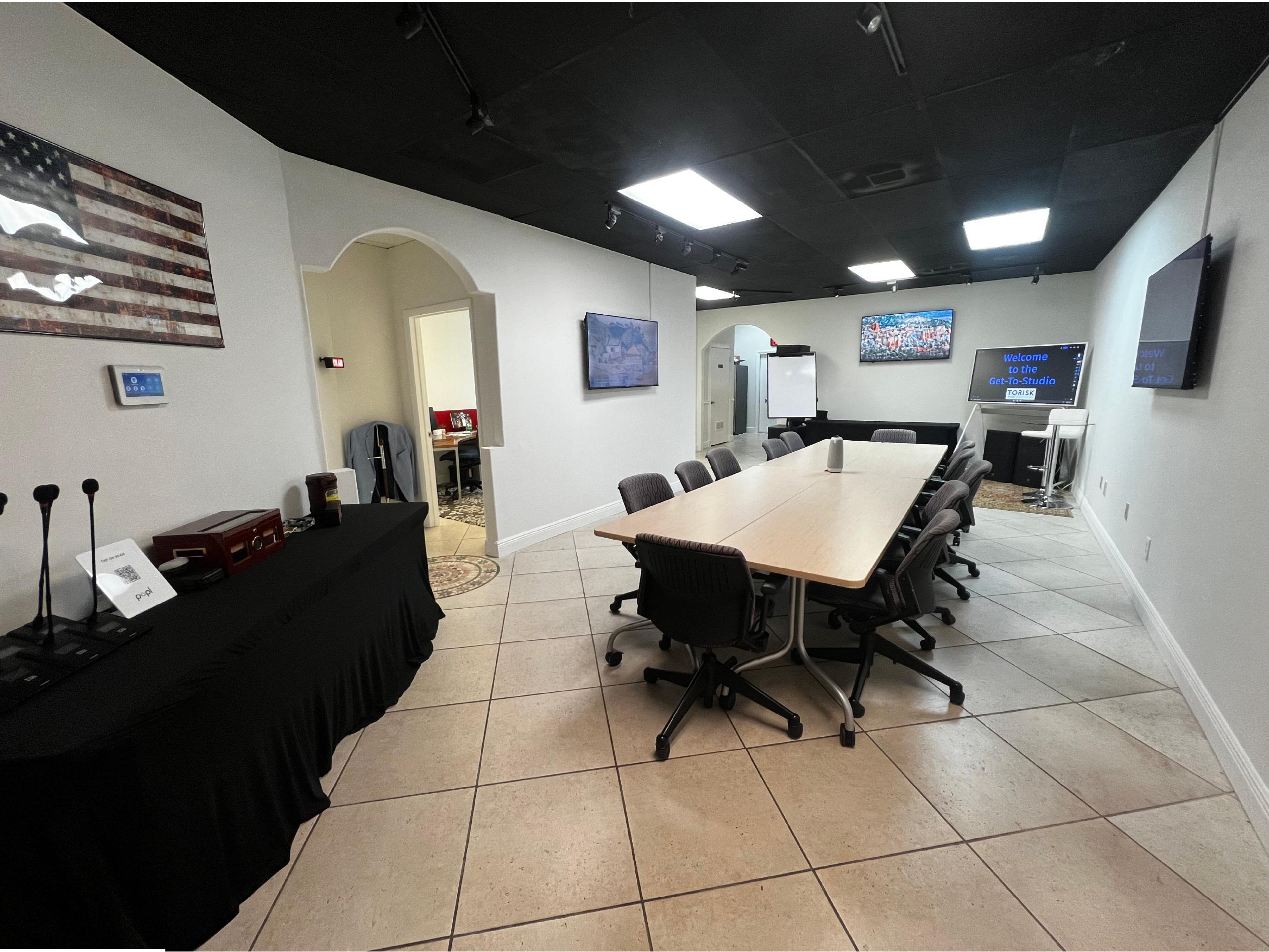The image shows a conference room with a long table surrounded by chairs, two large screens, and a side area with additional seating and a presentation board.