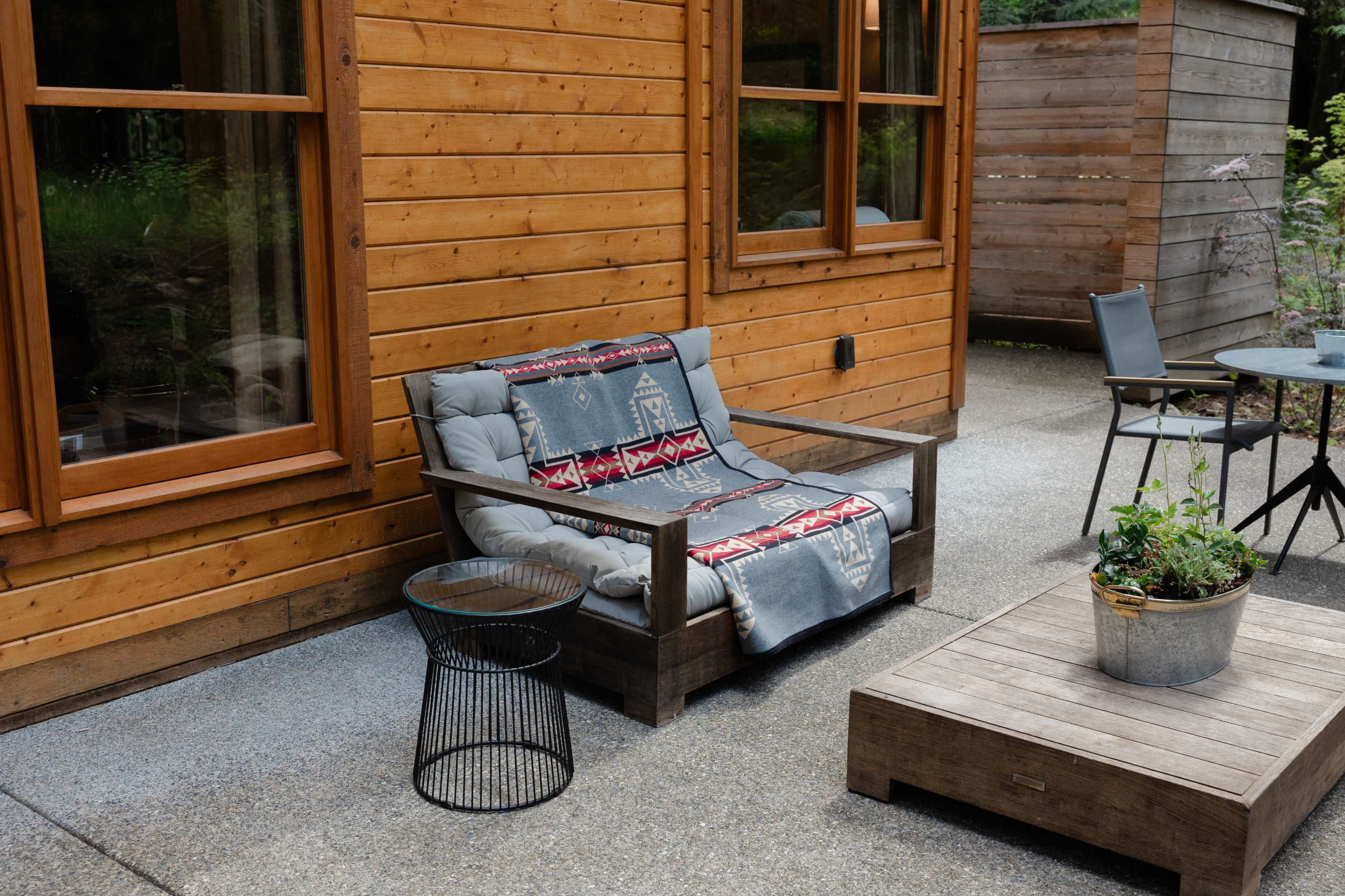 A wooden patio features a cushioned sofa covered with a blanket, a round table with a chair, and a planter on a concrete floor.