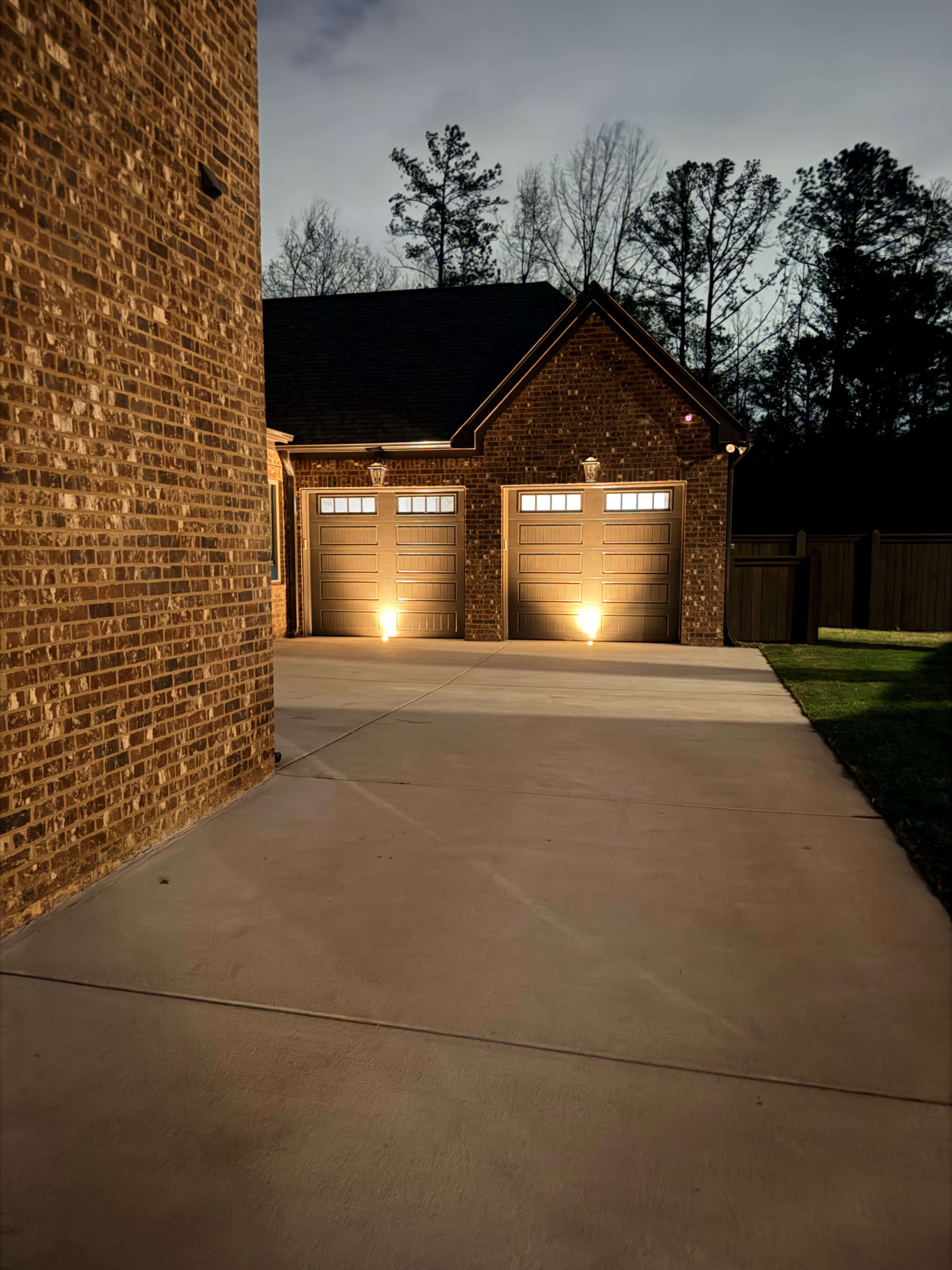 The image shows a brick house with two illuminated garage doors, set against a dark evening sky and surrounded by trees.