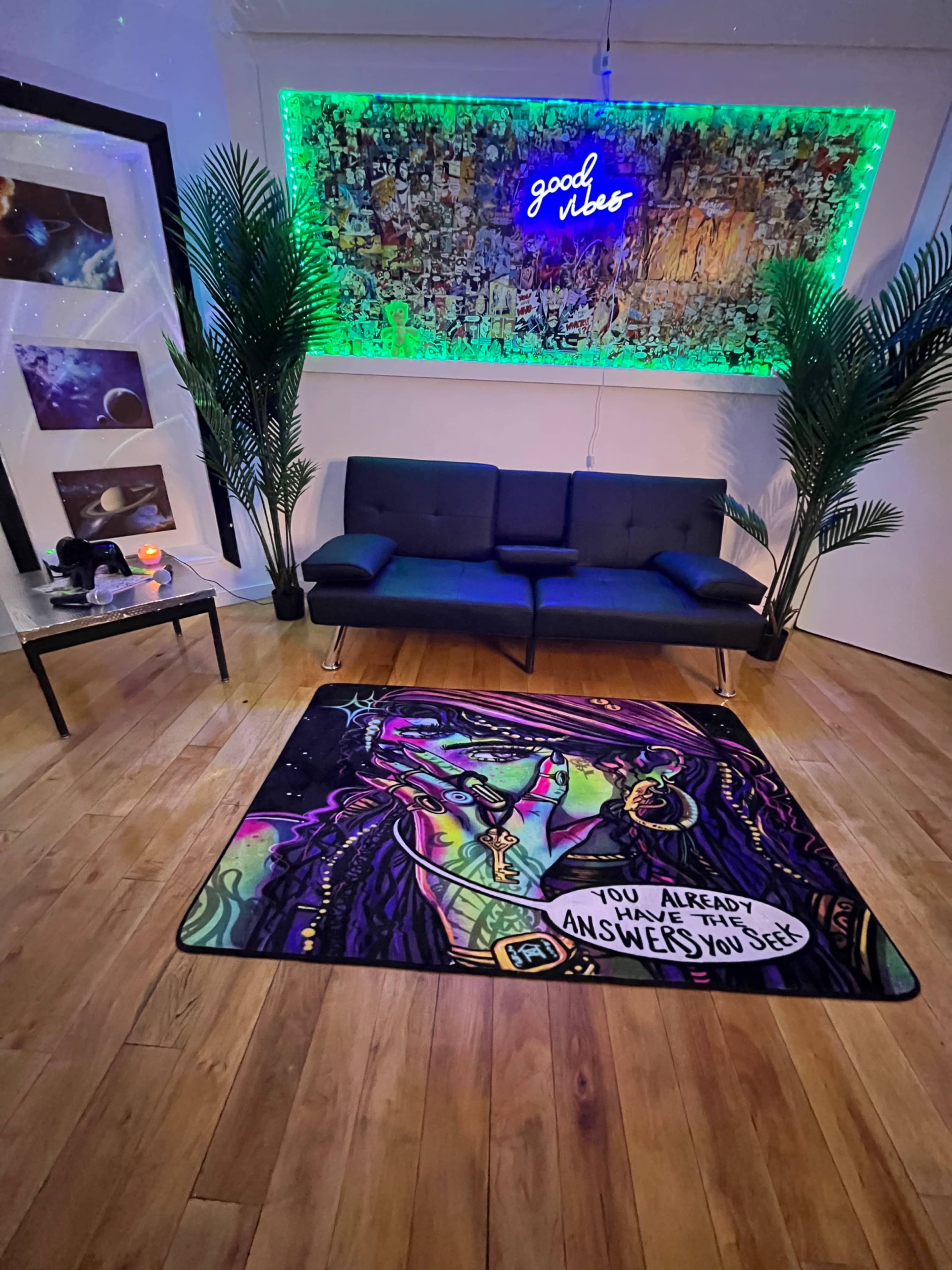 A colorful rug featuring a character with green hair and vibrant artwork is placed on a wooden floor in front of a black couch, framed art, and a neon sign that says "good vibes."