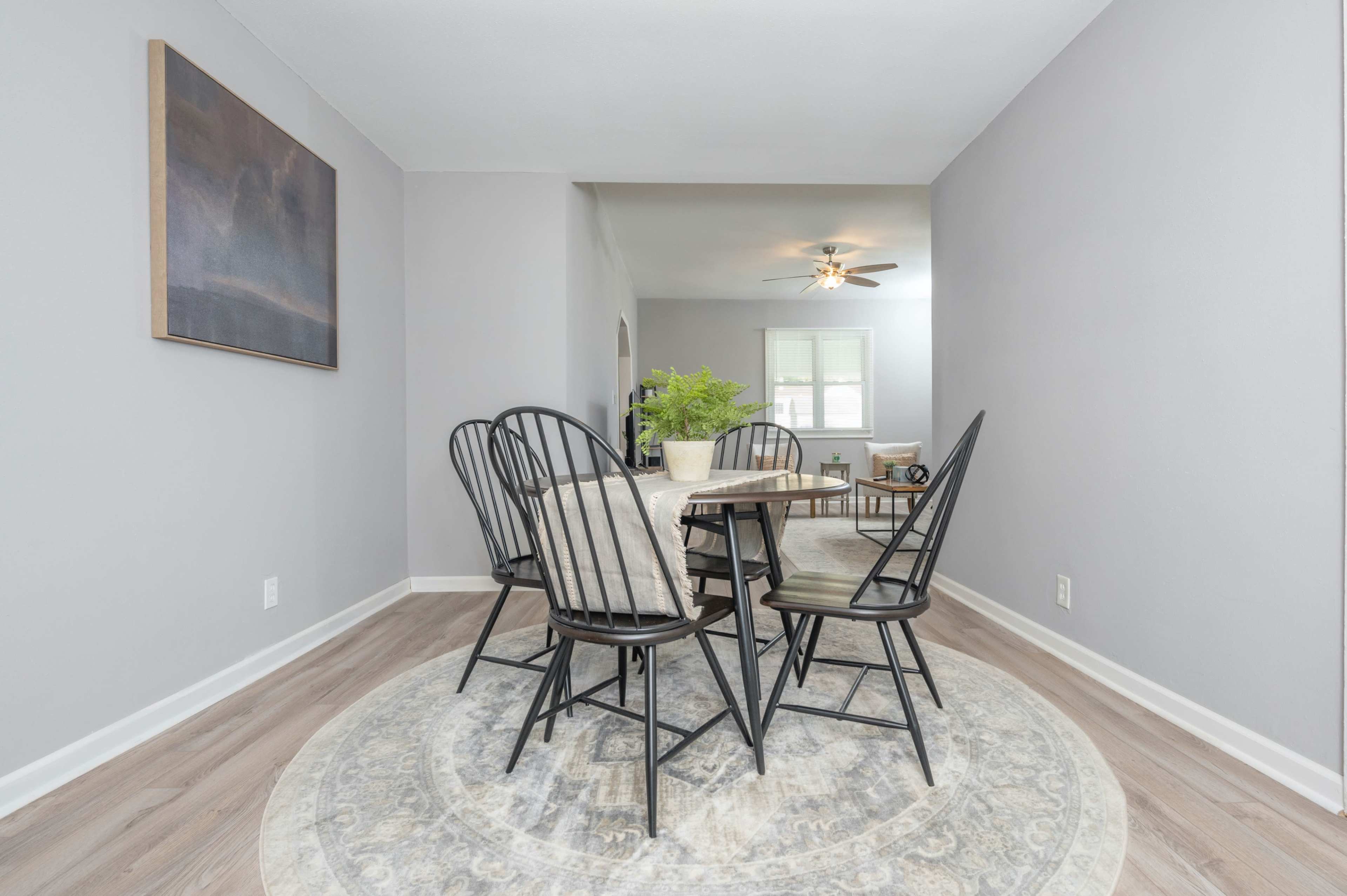 A dining area features a round table surrounded by black chairs on a patterned rug with gray walls and a ceiling fan above.