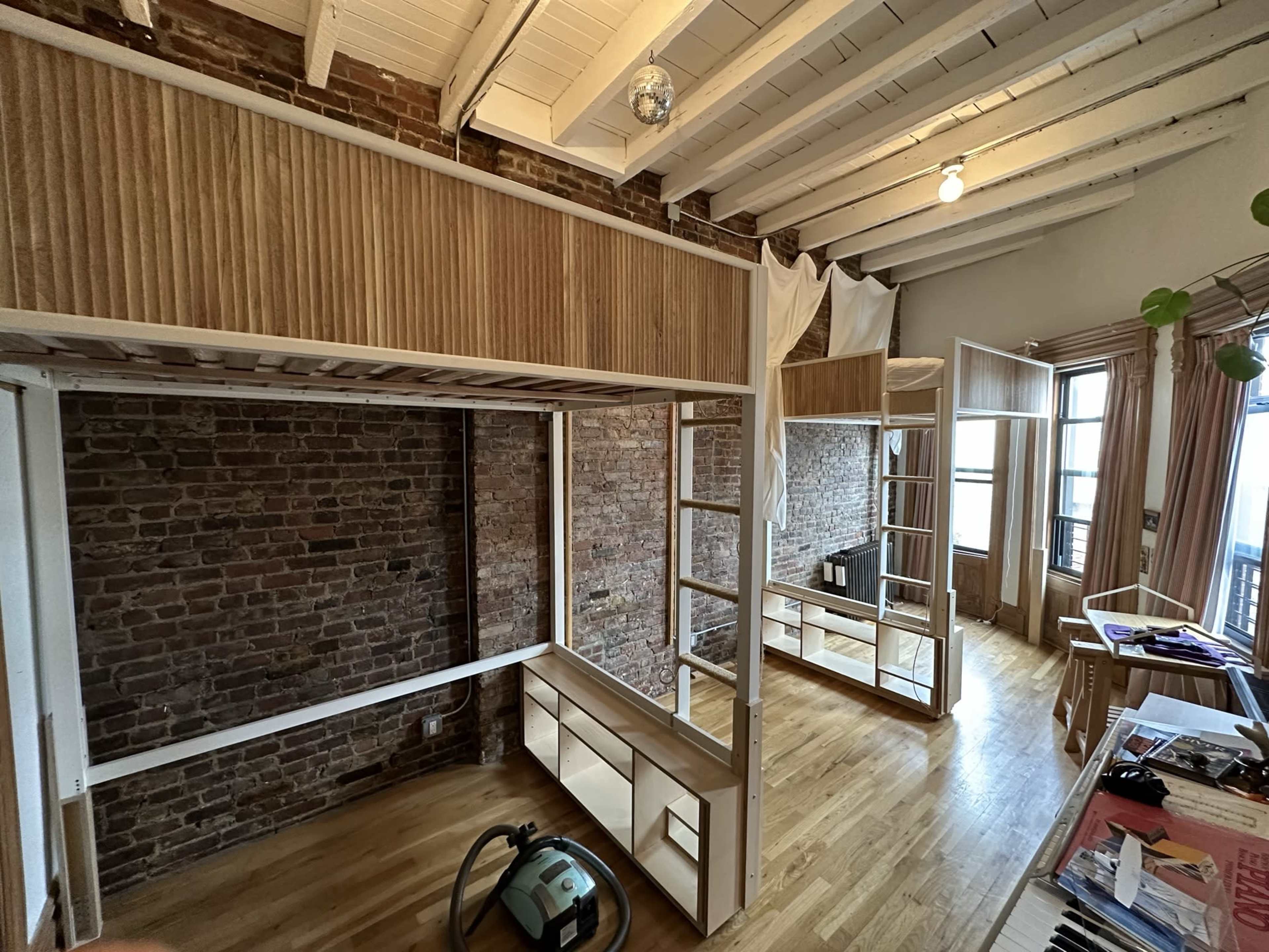 The image shows a spacious room with exposed brick walls, wooden beams, and built-in shelving units, featuring a vacuum cleaner on the floor.
