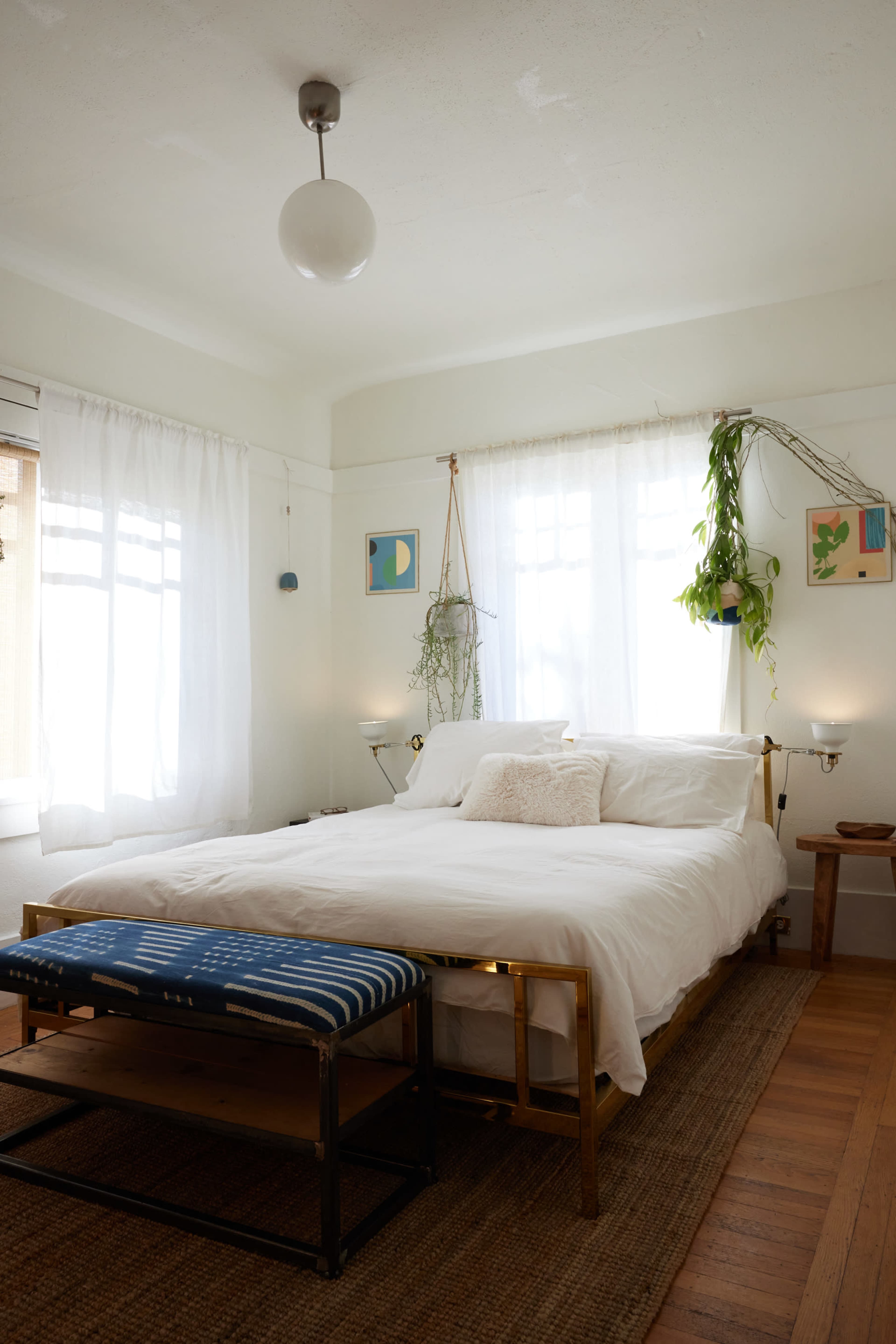 A neatly arranged bedroom features a large white bed with a decorative throw, a woven rug, plants hanging by the windows, and framed artwork on the walls.