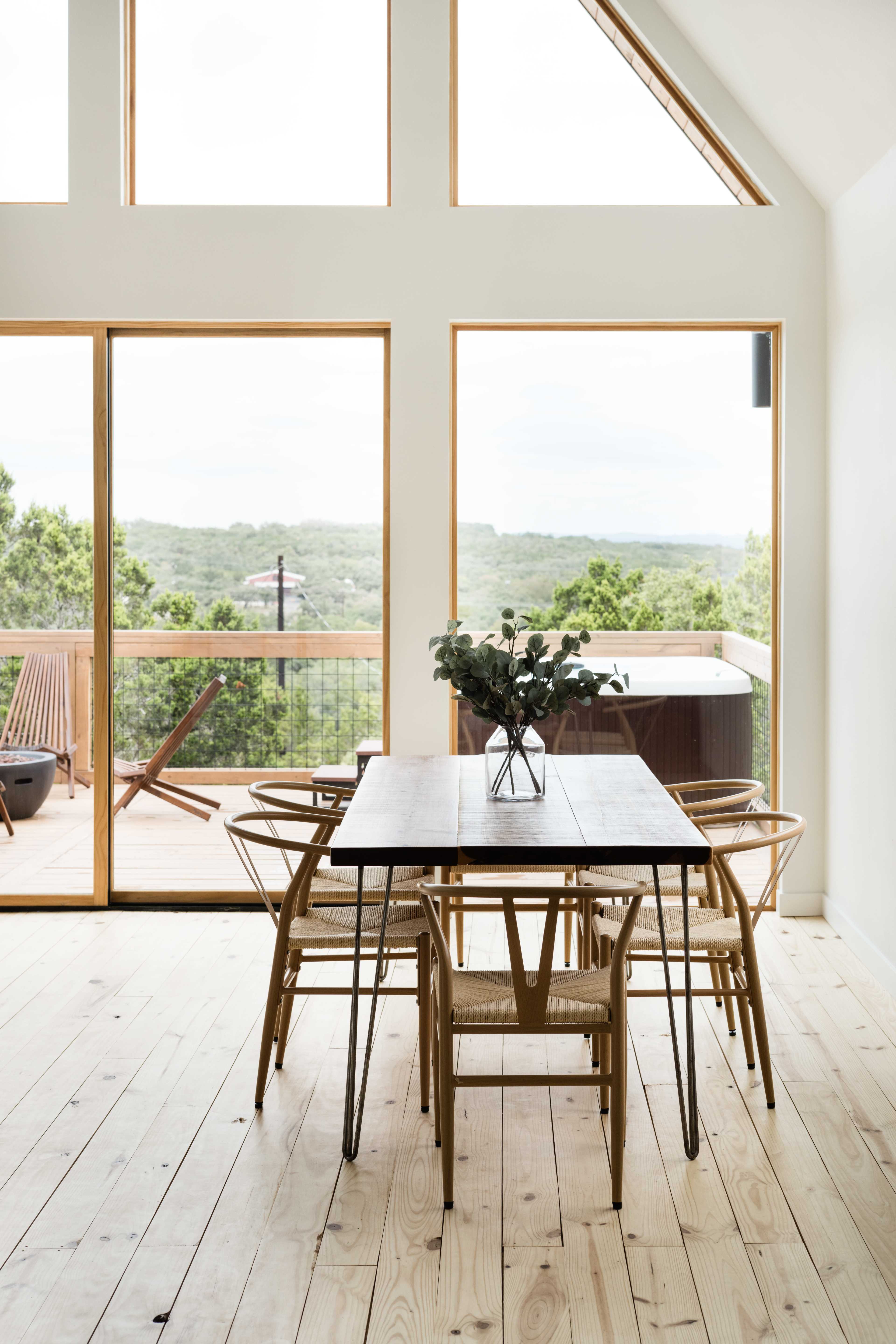 A modern dining area features a wooden table surrounded by chairs, with large windows offering a view of the outdoors and a deck with a hot tub.