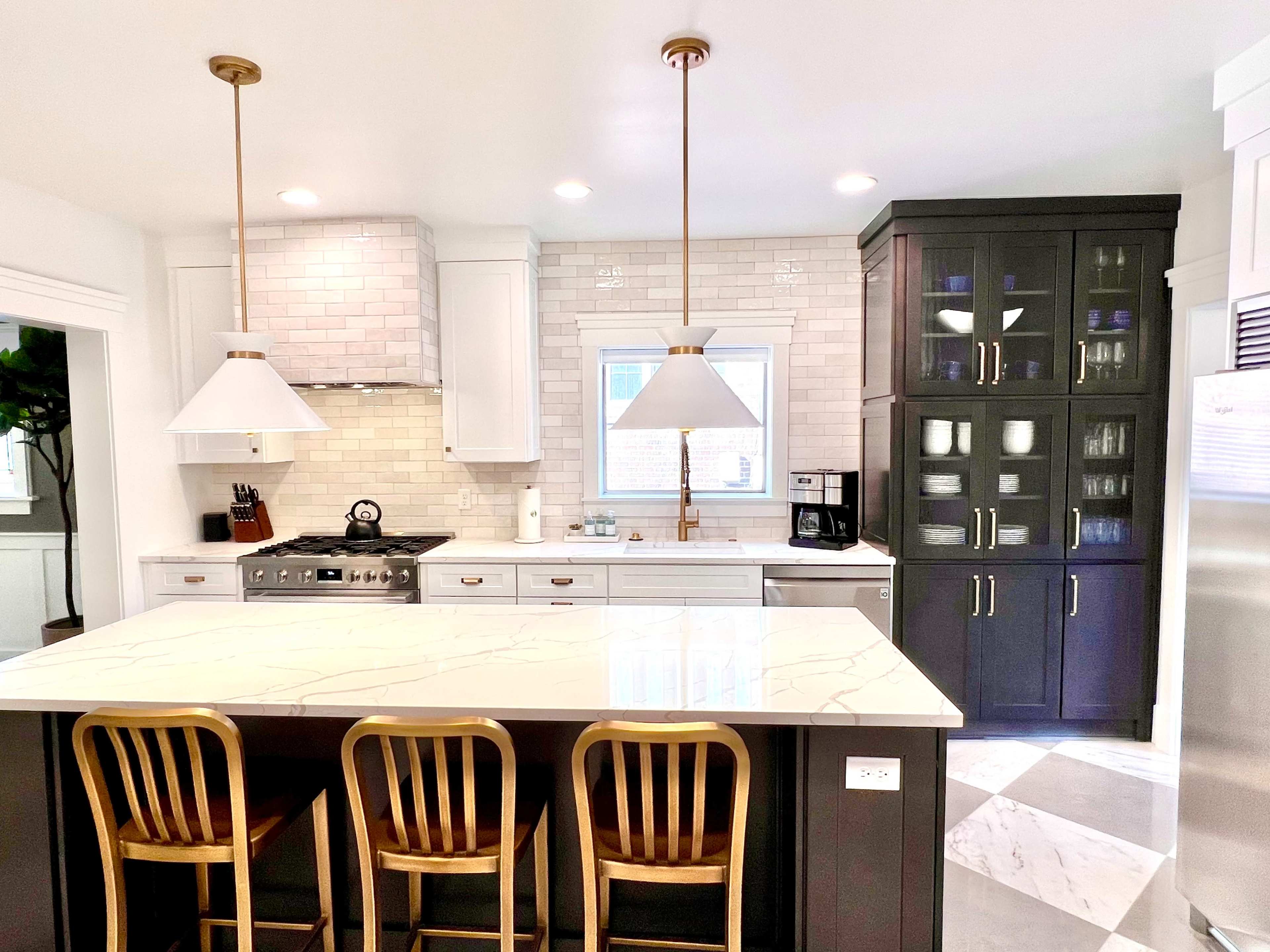 A modern kitchen features a central island with three bar stools, pendant lights above, and a combination of dark cabinetry and white tile backsplash.