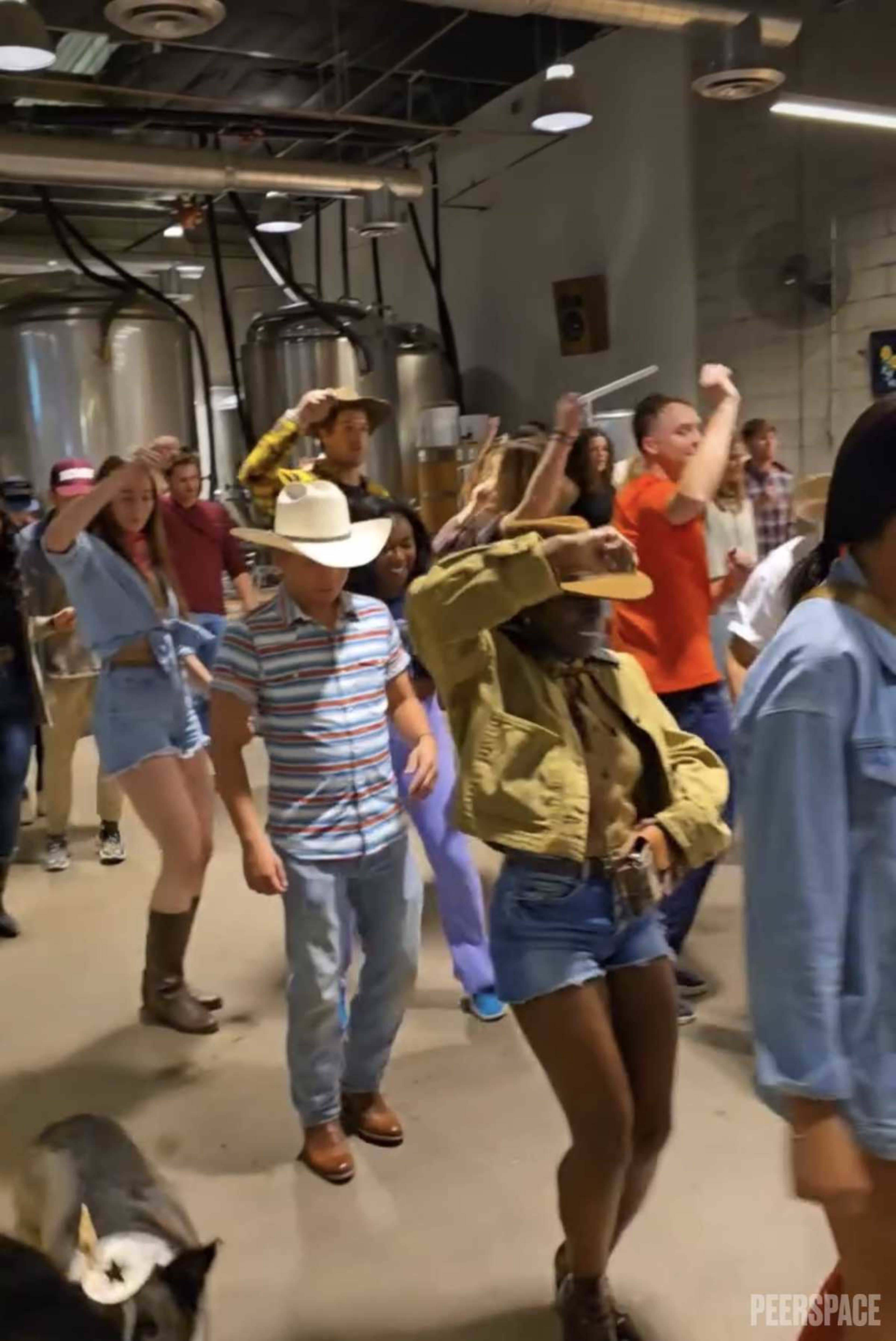 A group of people is line dancing inside a venue with large brewing tanks in the background.