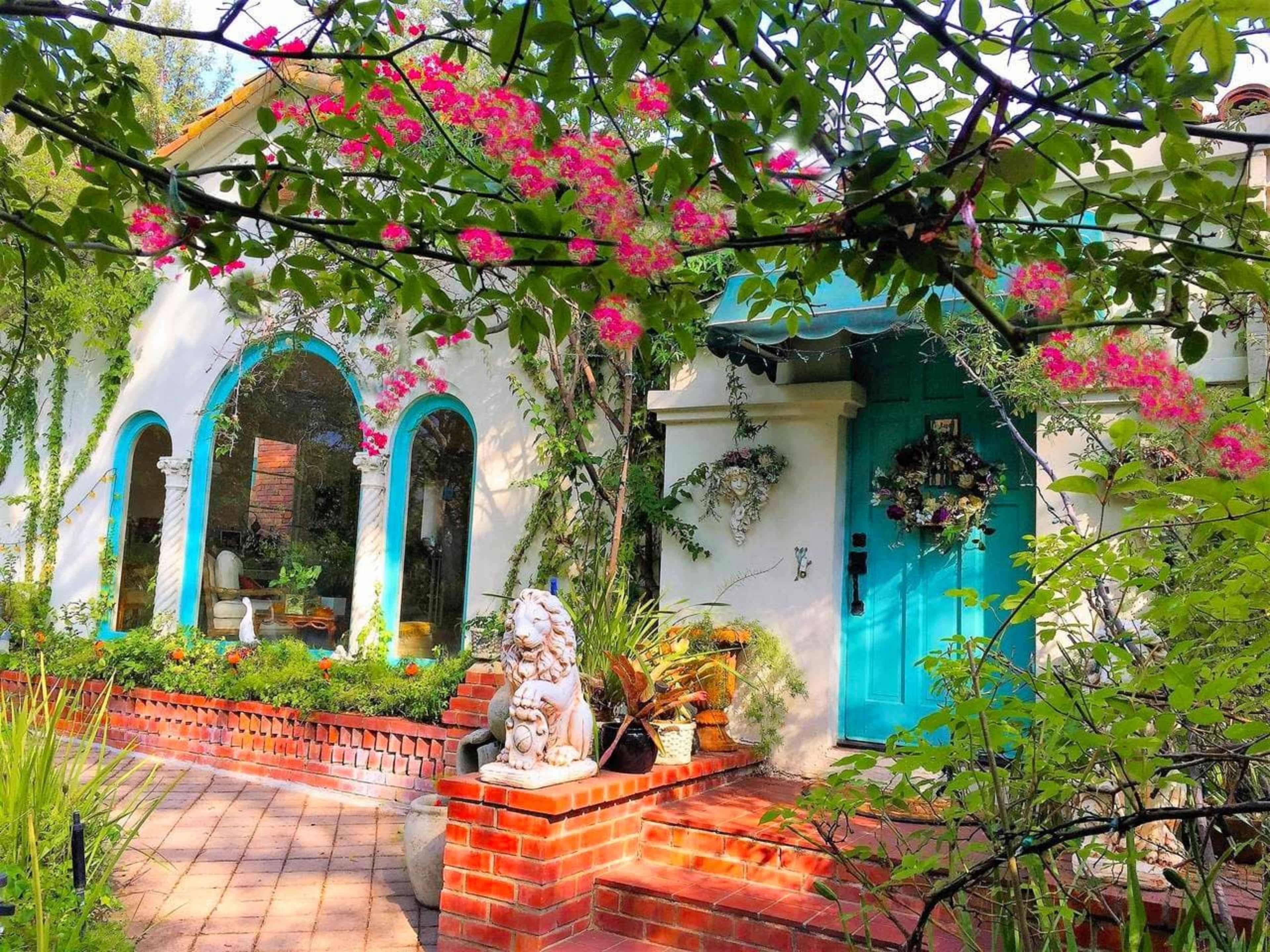 A colorful house with arched windows and a turquoise door is surrounded by lush greenery and blooming flowers.