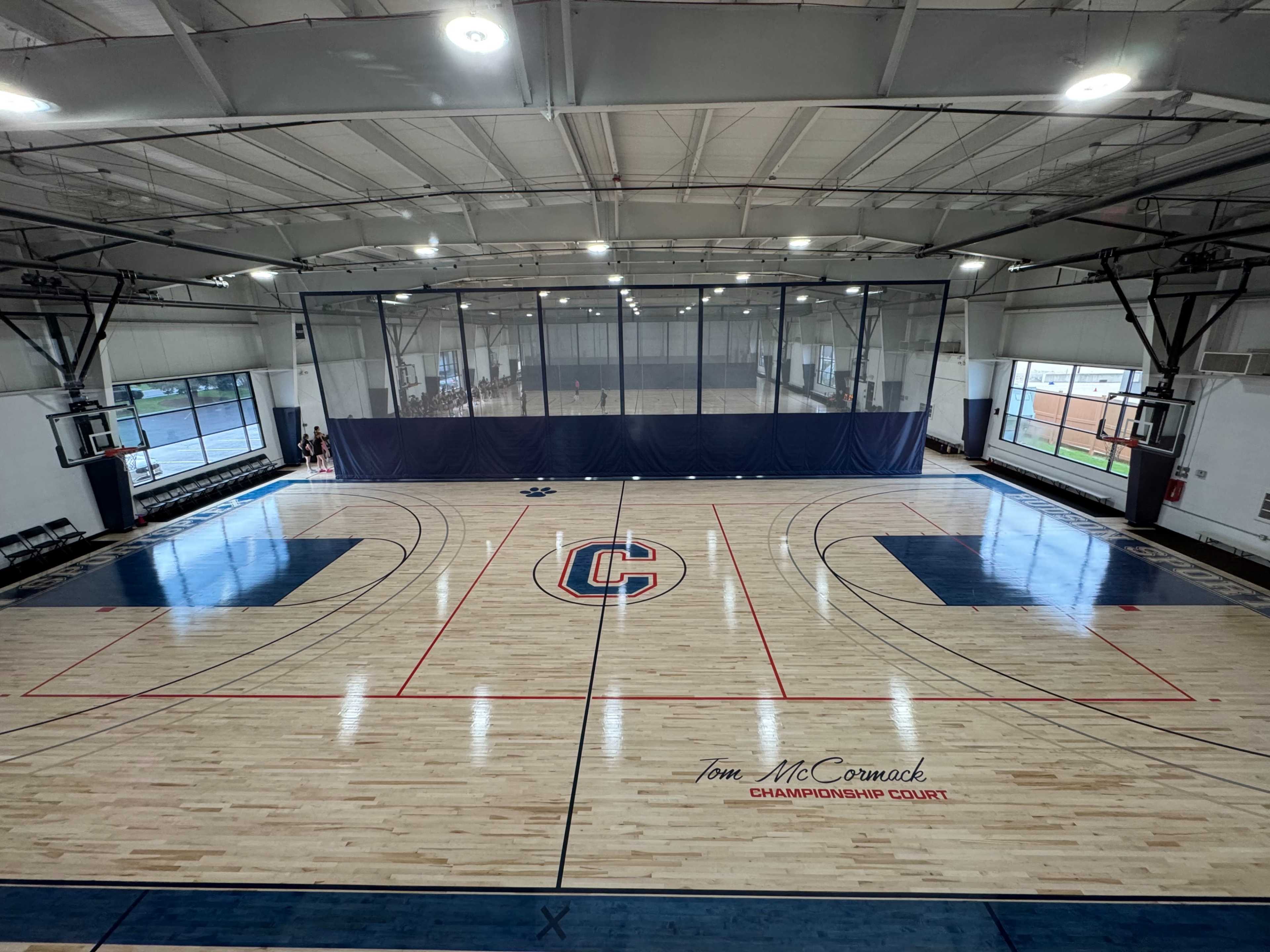 The image shows a spacious indoor basketball court with a polished wooden floor, featuring marked lines and a large logo at the center.