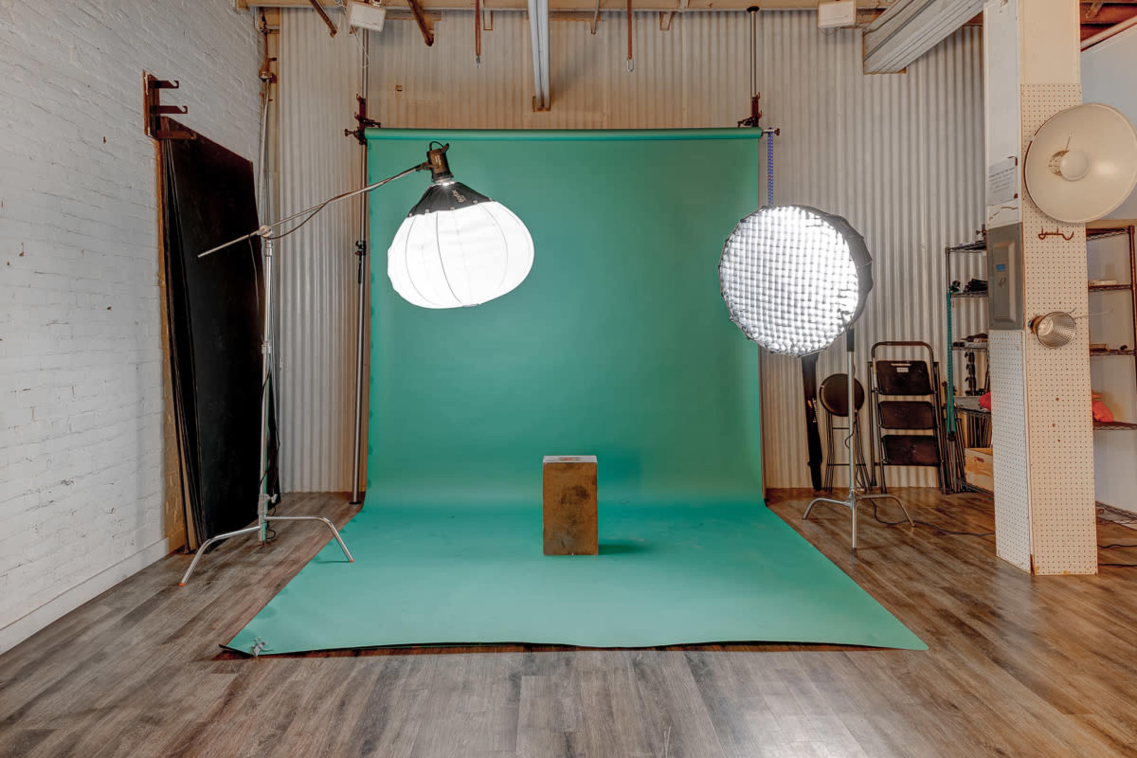 The image shows a photography studio setup with a green backdrop, a stool, and two lighting sources positioned on either side.