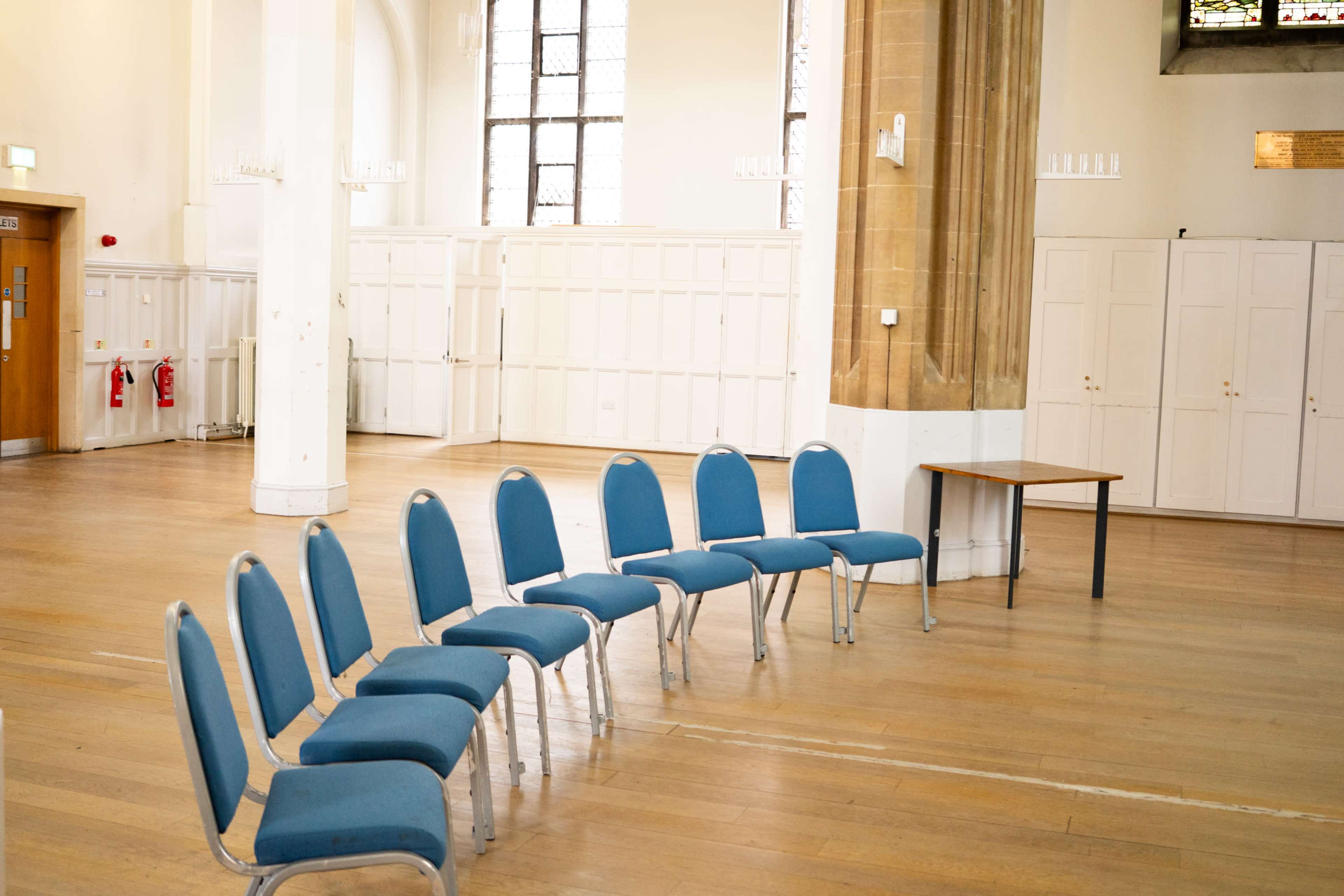 A semicircle of blue chairs is arranged next to a wooden table in a spacious interior with wooden floors and large windows.