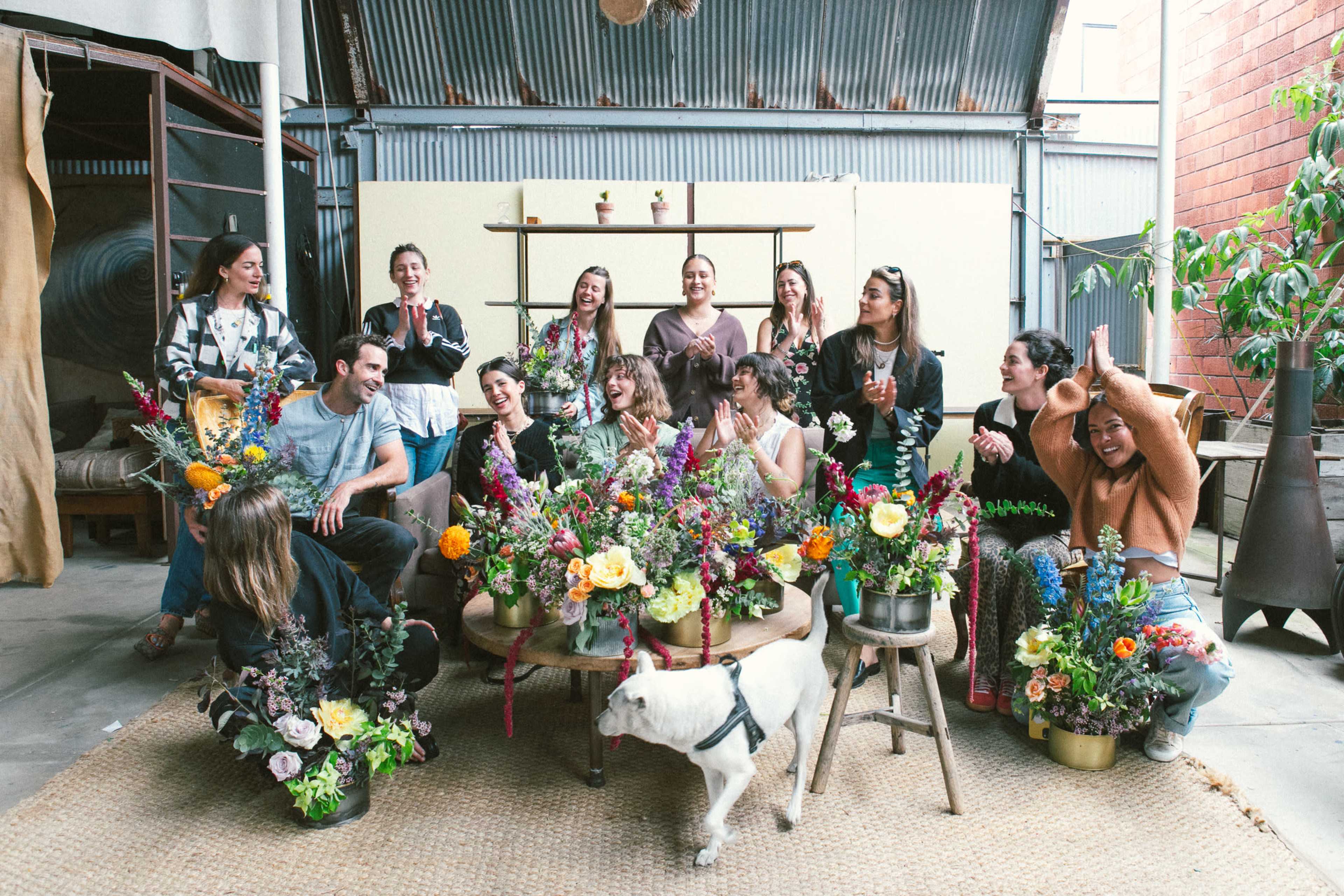 A group of people is gathered in a decorated indoor space filled with flowers, with a white dog sitting in the front.
