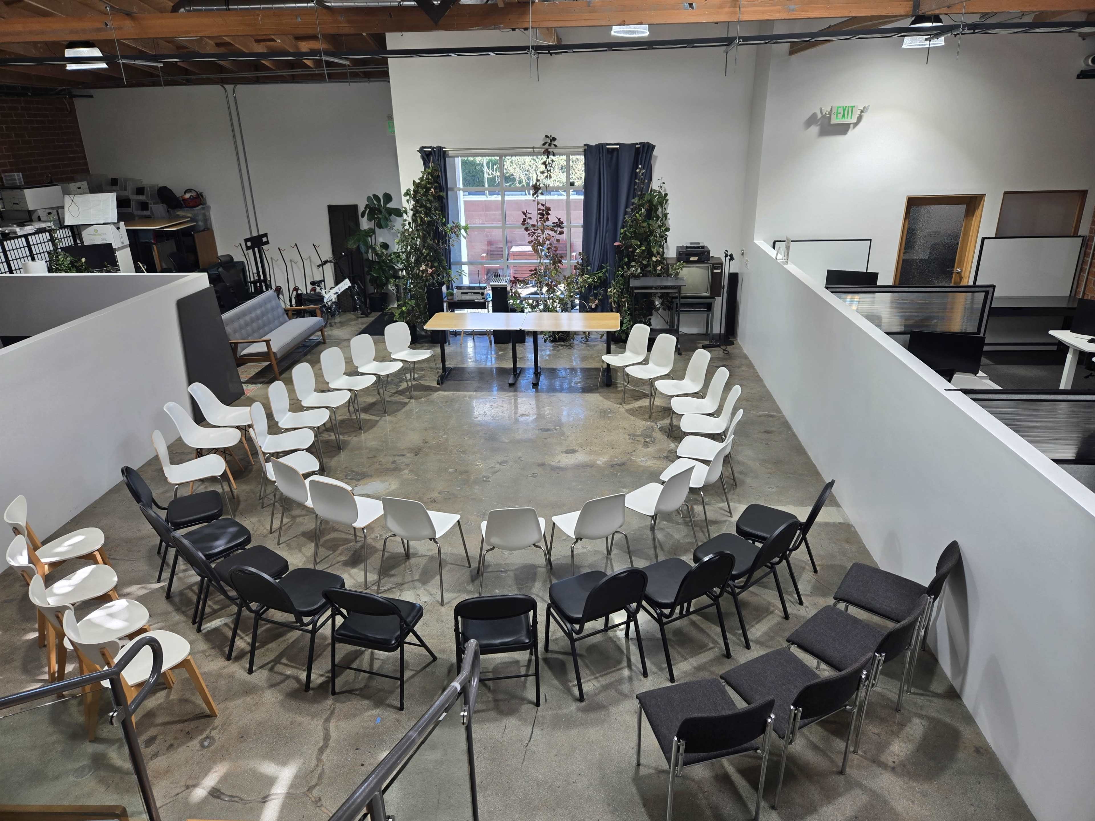 The image shows a circular arrangement of chairs around a central table in a bright, open meeting space with plants and natural light.