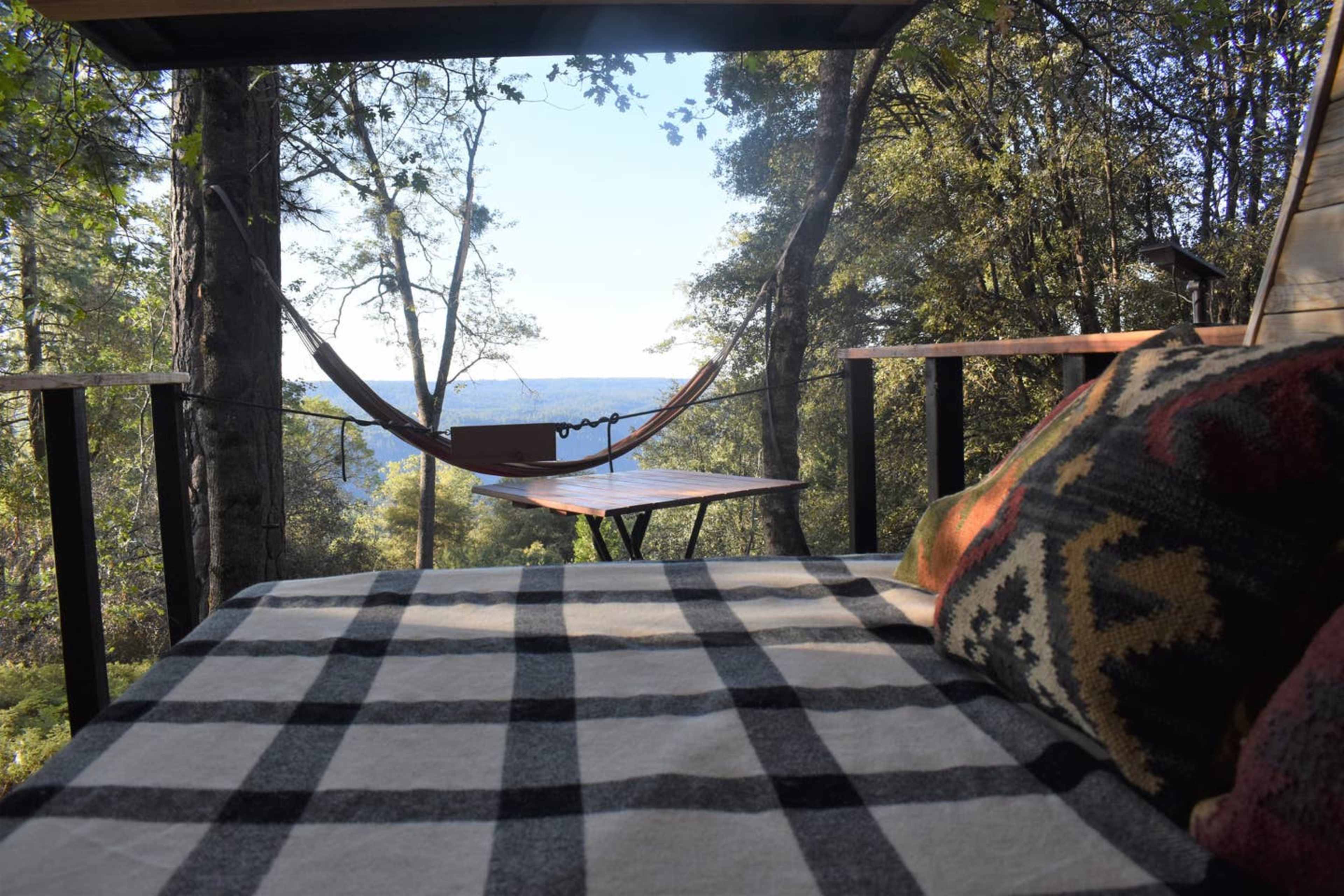 A cozy treehouse interior features a bed with a patterned blanket, a small table, and a hammock overlooking a scenic view through the trees.