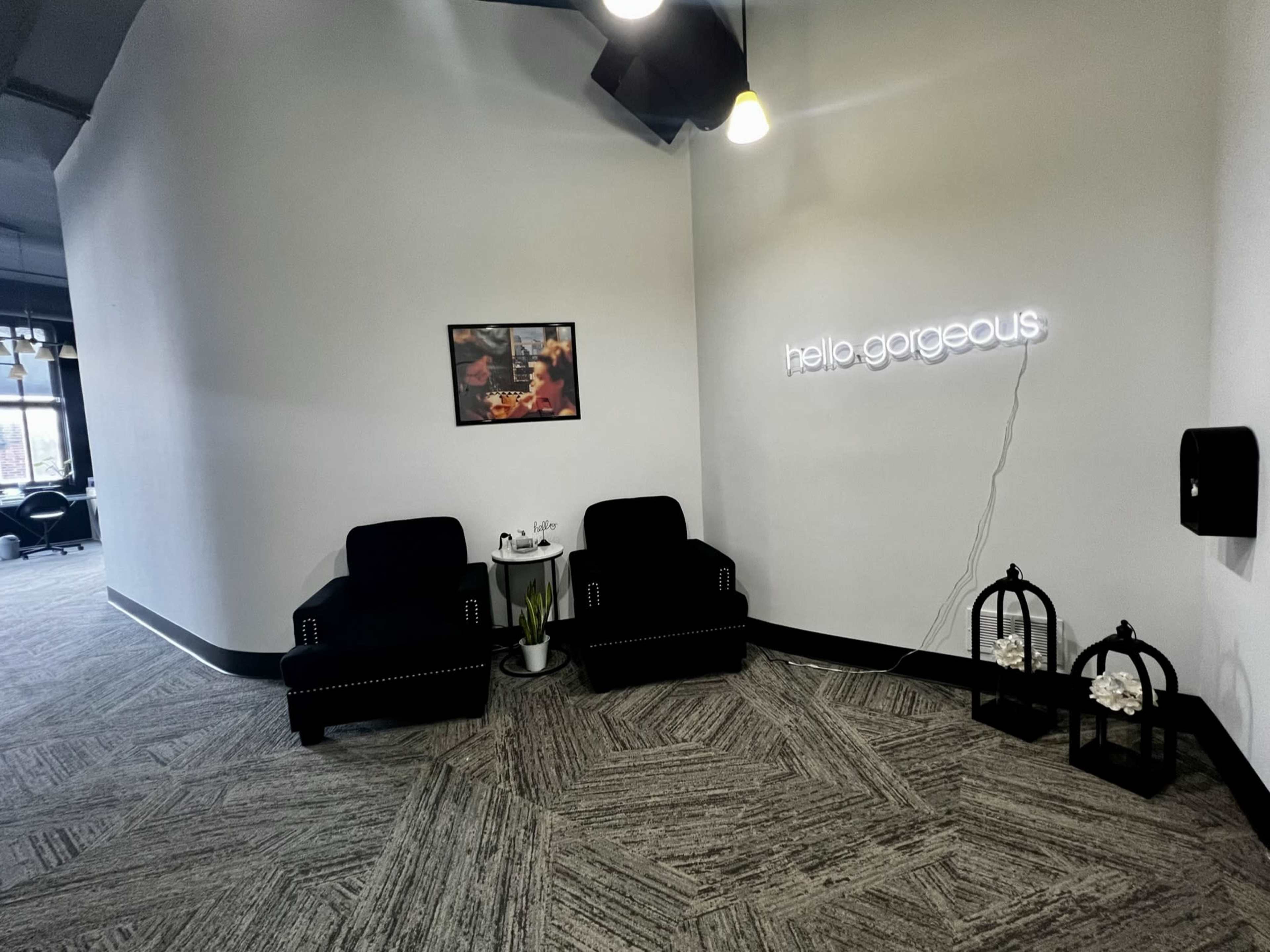 A modern indoor waiting area featuring two black armchairs, a framed photo on the wall, and a neon sign that reads "hello, gorgeous."