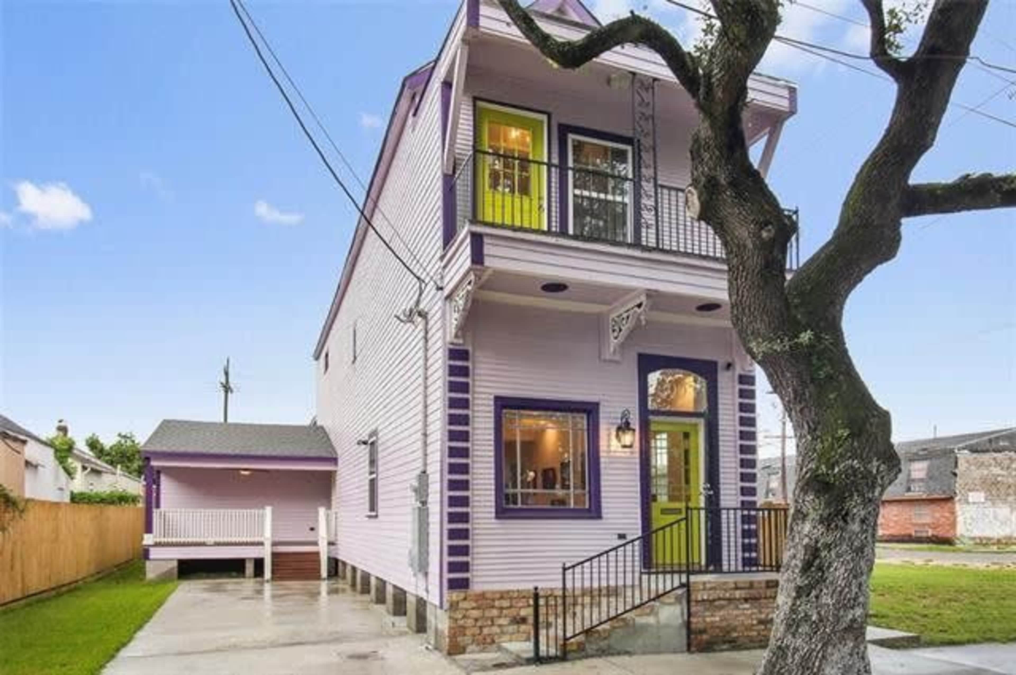 A three-story, purple and white house with green accents, featuring a porch and a tree in the front yard.