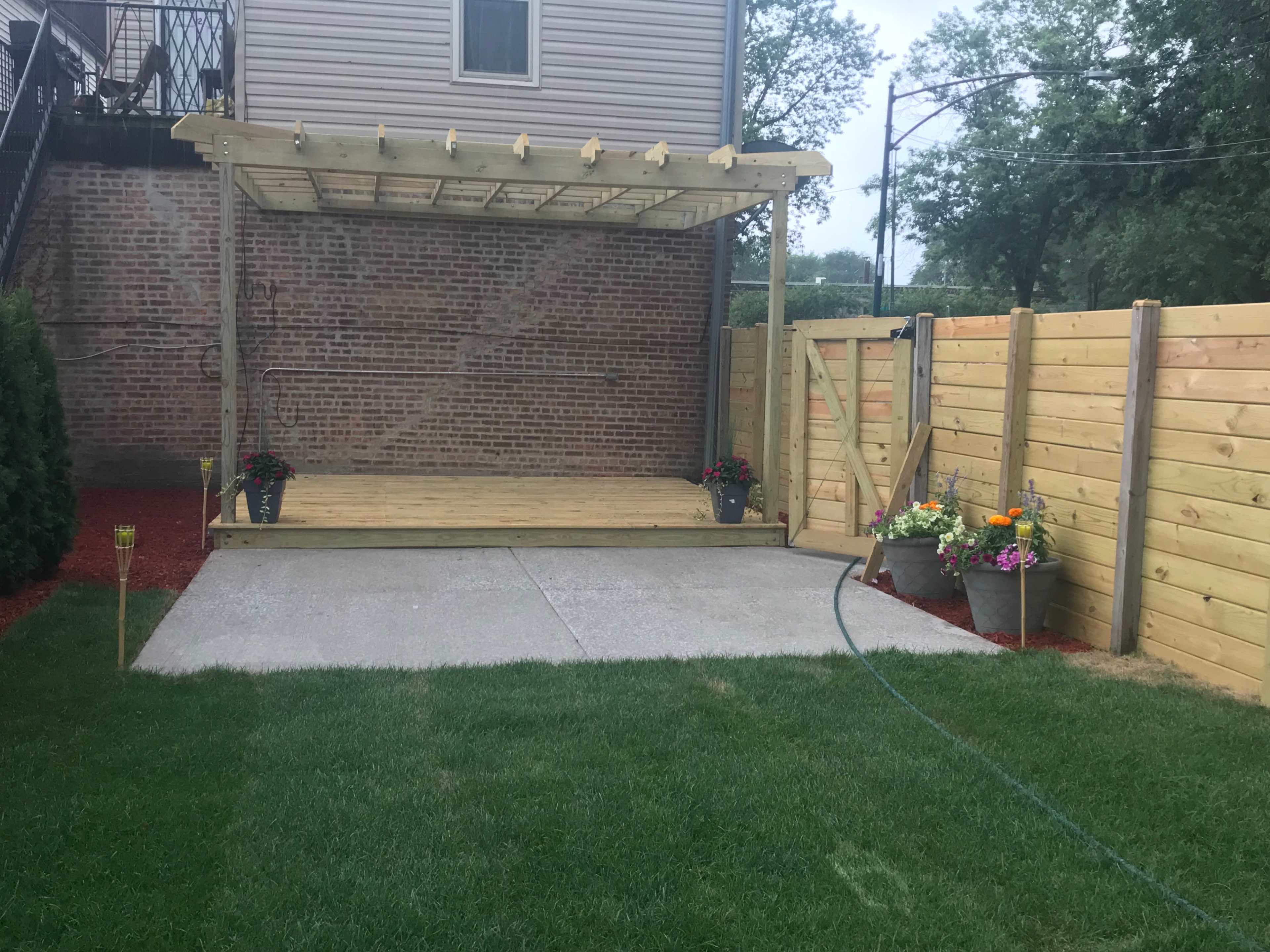 A wooden deck with a pergola, surrounded by a fenced yard with flower pots and a grassy area.