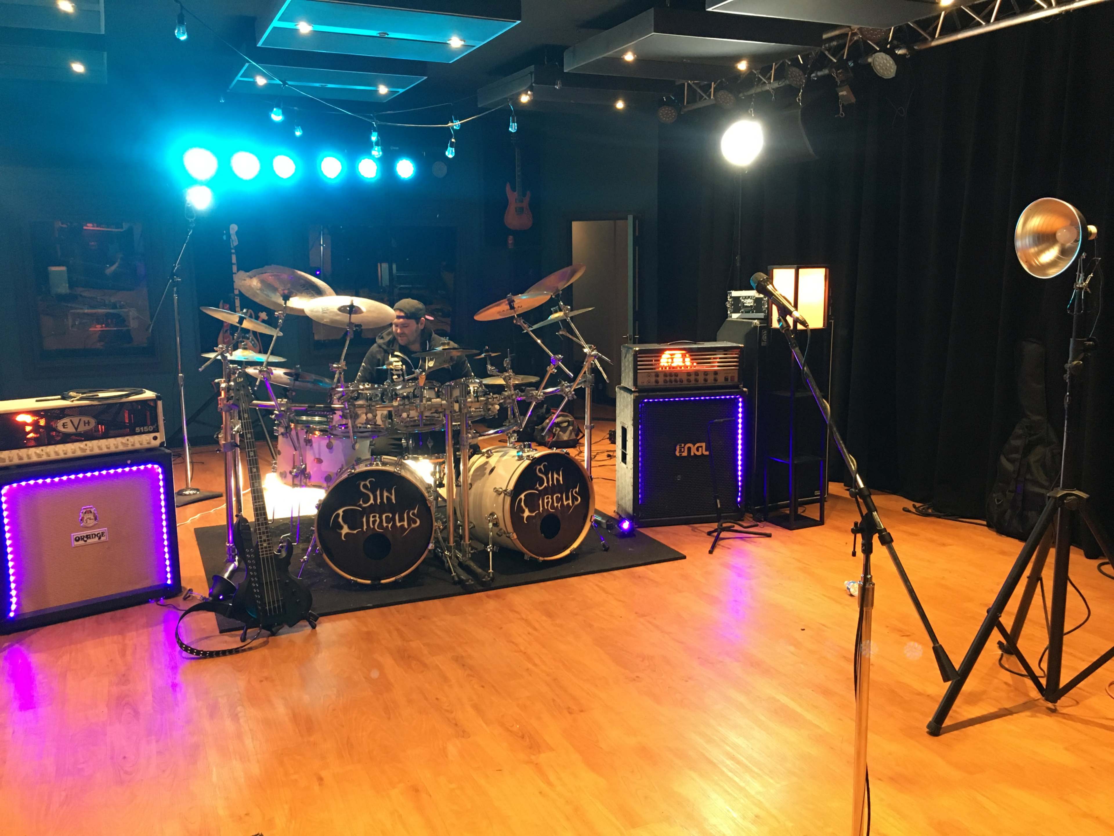 A drummer sits at a drum set labeled "Sin Circus" in a music studio equipped with amplifiers and colorful stage lights.