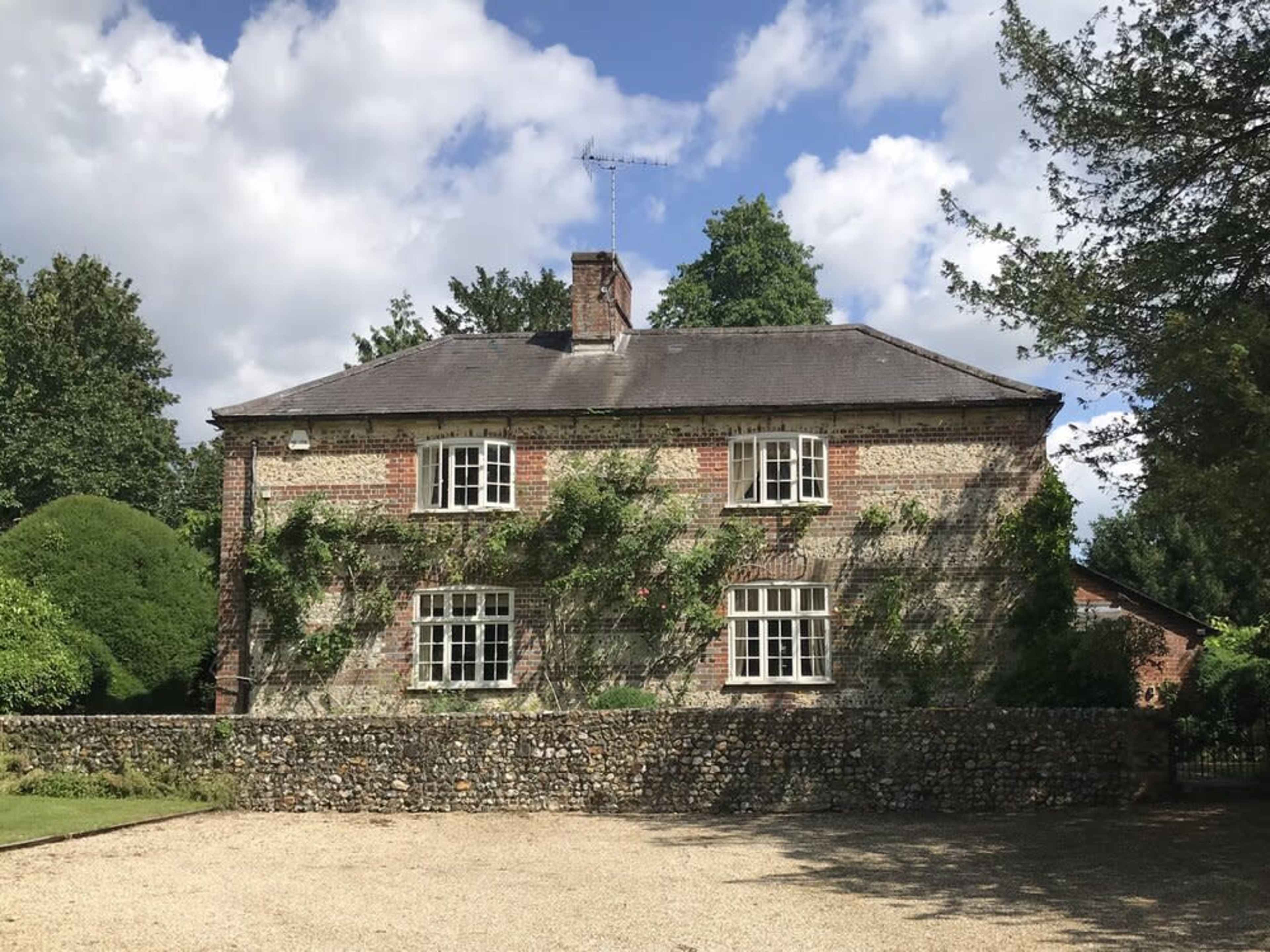 A two-story brick house with climbing plants and multiple windows is set behind a stone wall, surrounded by greenery under a partly cloudy sky.